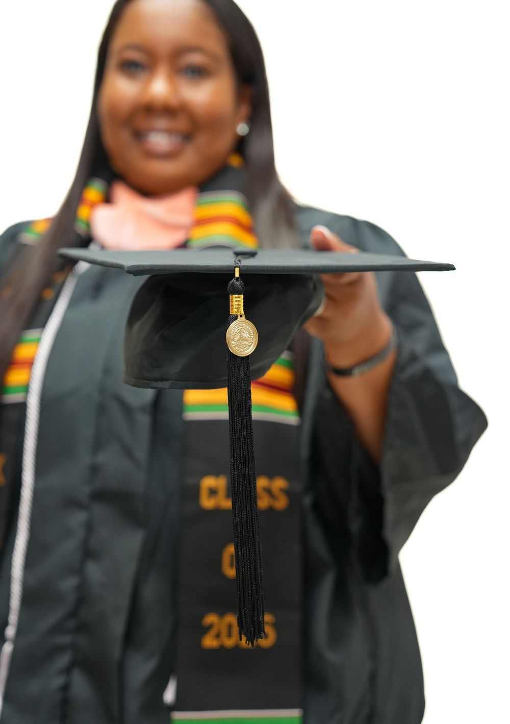 Smiling graduate in dark green cap and gown holding mortarboard forward, wearing kente cloth stole, Class of 2025.