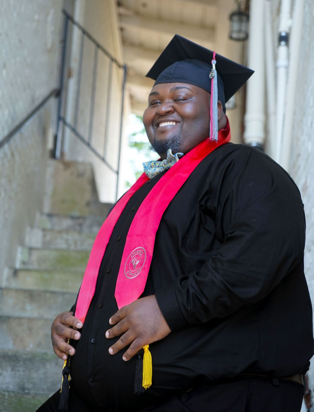 Smiling graduate in black cap and gown with red stole poses proudly on steps at outdoor graduation ceremony.