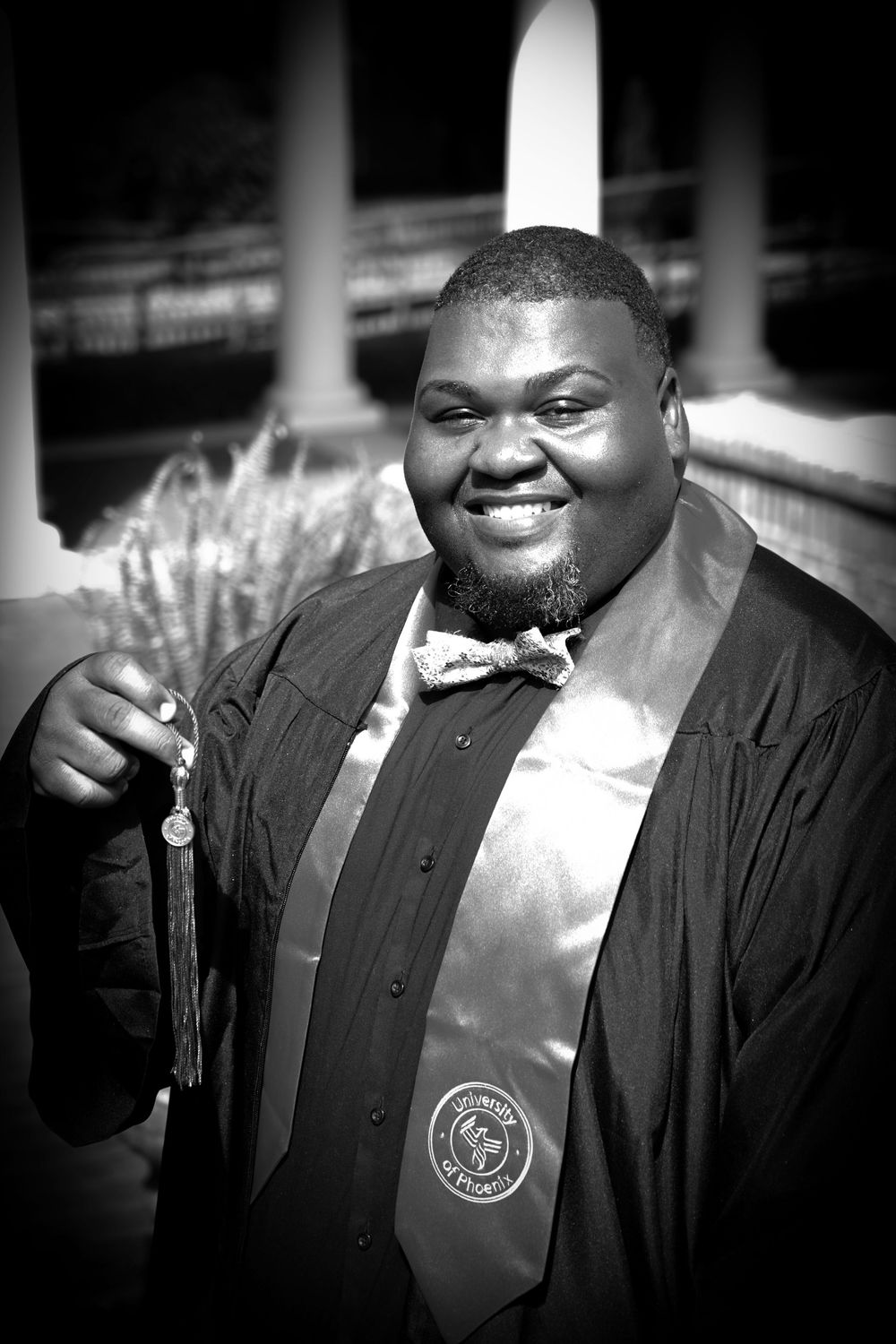 Smiling graduate in cap and gown holding tassel, wearing bow tie and academic stole, black and white photo.