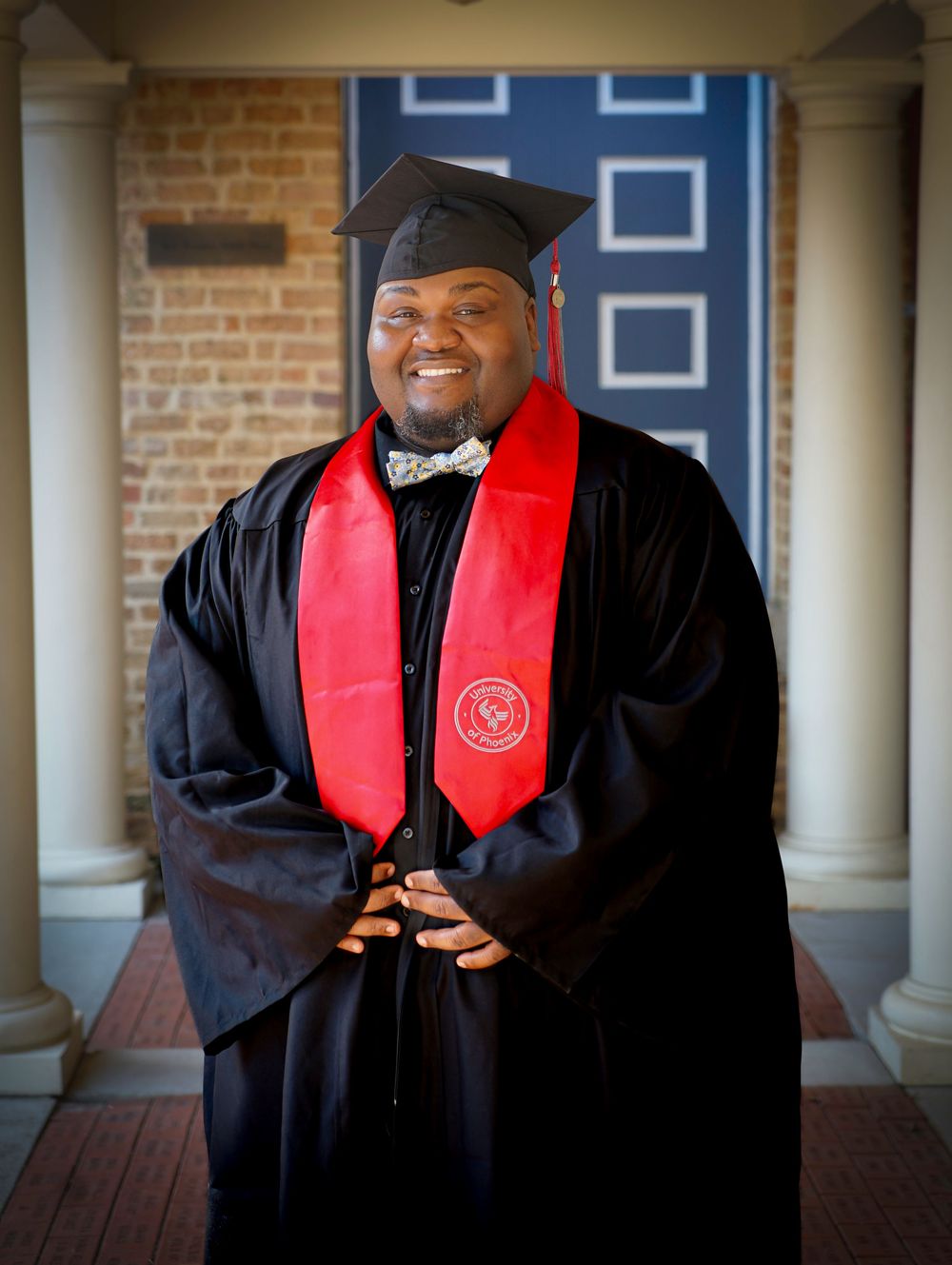 Smiling graduate in black cap and gown with red honor stole poses proudly in front of a brick building.