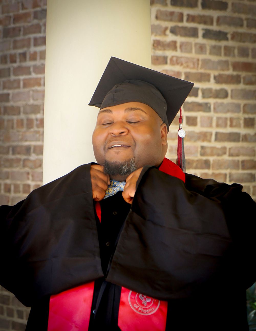 A proud graduate in black cap and gown with red sash smiles joyfully during graduation ceremony near brick wall.
