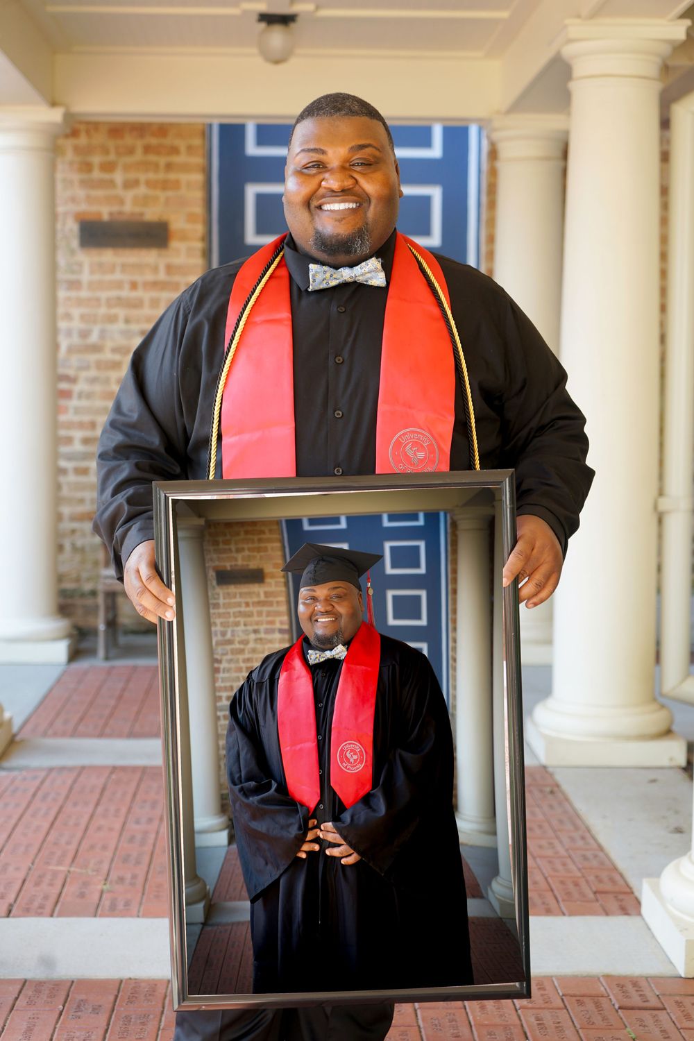 Man in graduation attire holds a framed photo of himself in cap and gown, celebrating his academic achievement.