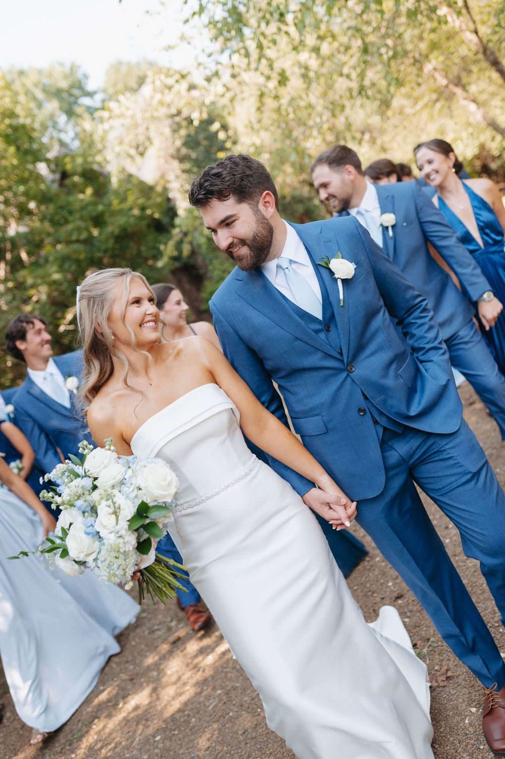 Bride and groom hold hands smiling with wedding party in blue attire outdoors.
