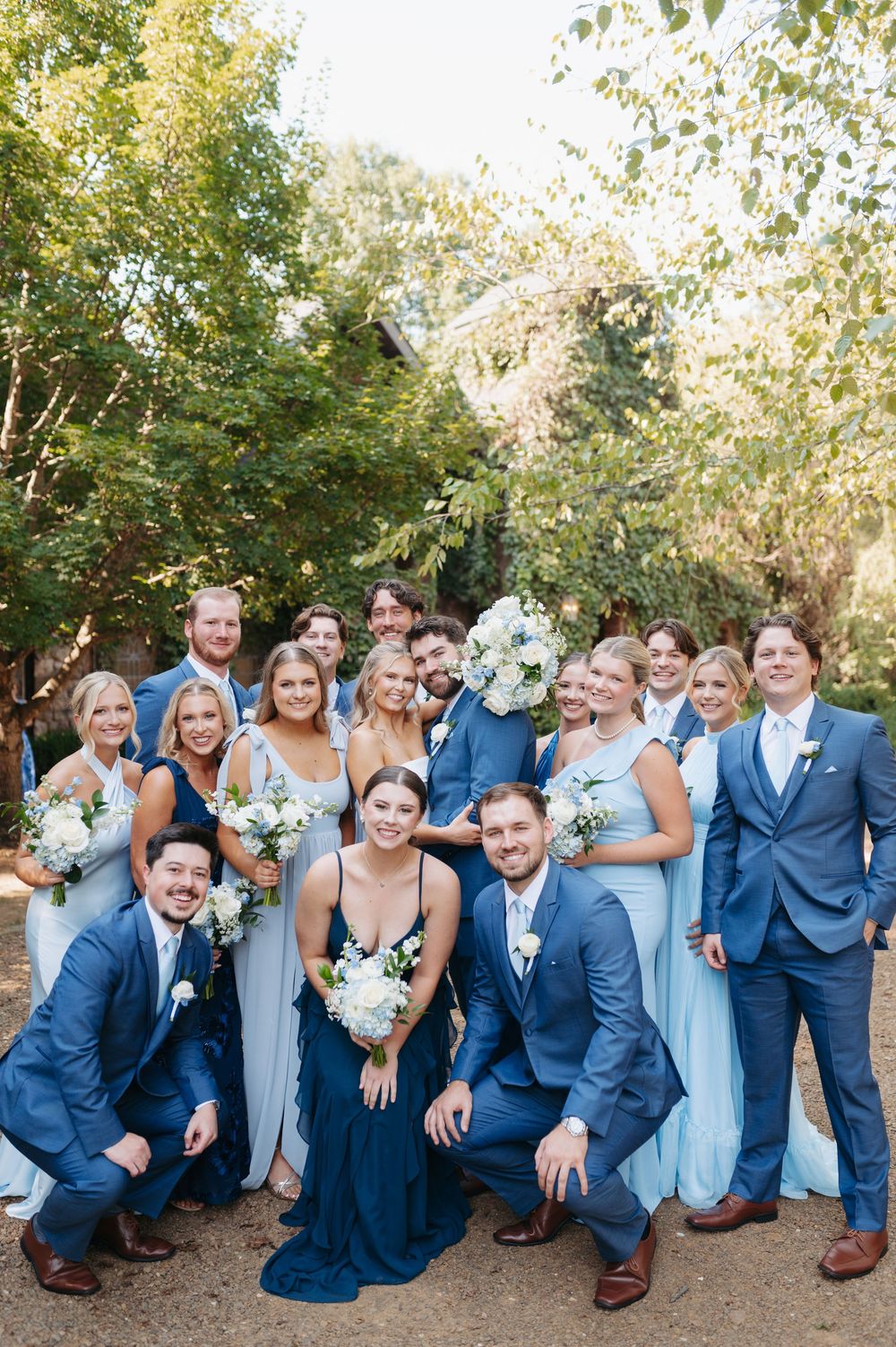 Wedding party in blue suits and light blue gowns poses outdoors with white bouquets among lush green trees.