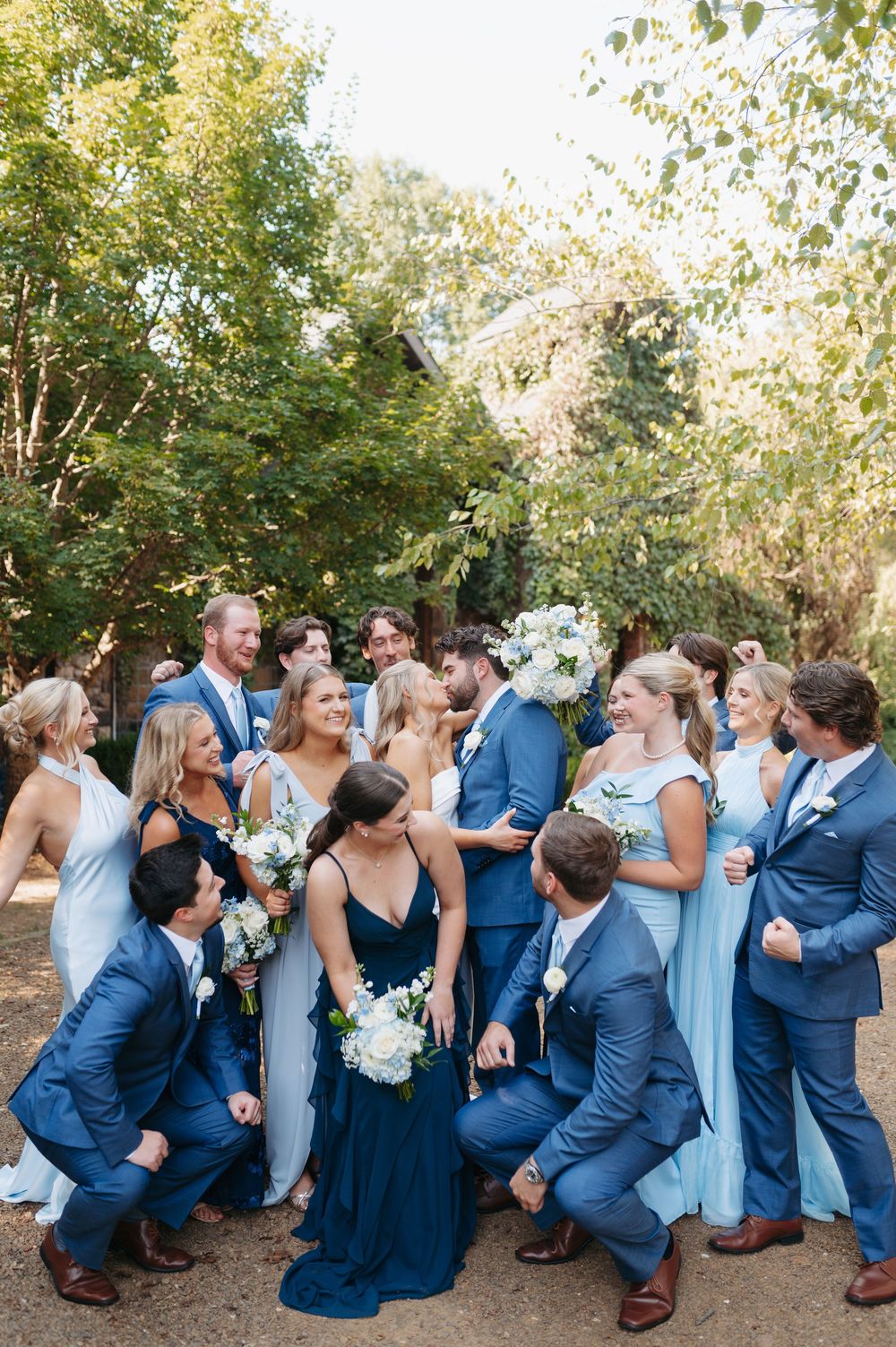 Joyful wedding party in blue suits and mismatched blue bridesmaid dresses celebrating outdoors with white floral bouquets.