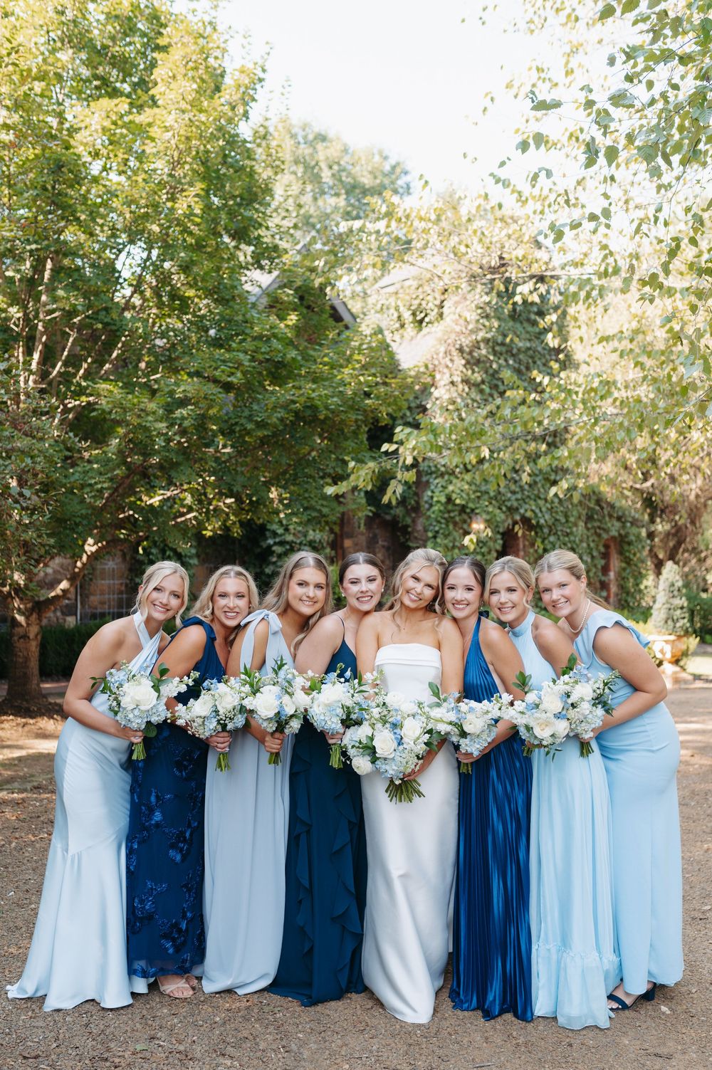 Bride in white gown poses with seven bridesmaids in blue dresses holding white bouquets outdoors.