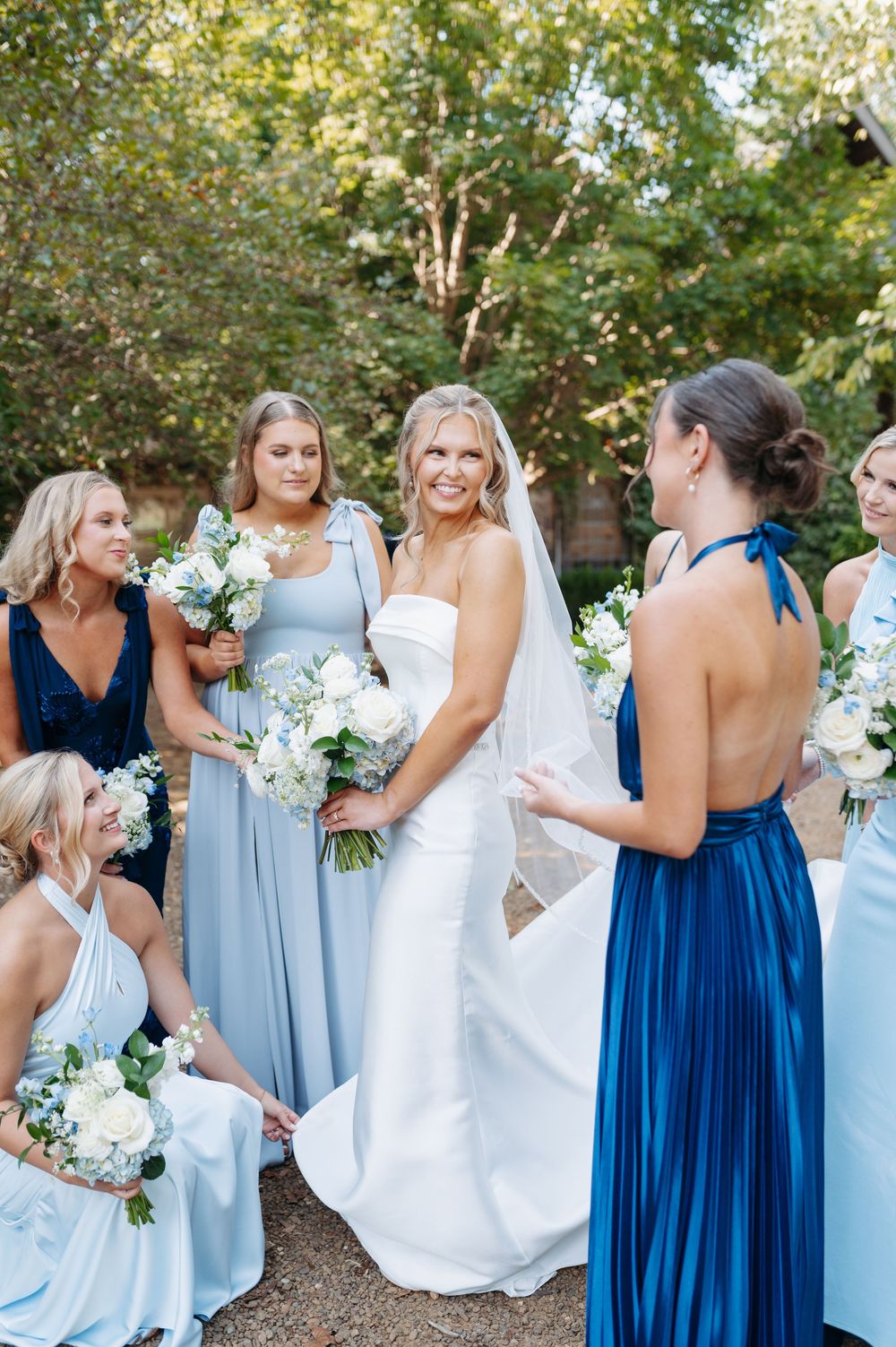 Bride in white gown with bridesmaids in blue dresses holding white floral bouquets outdoors.