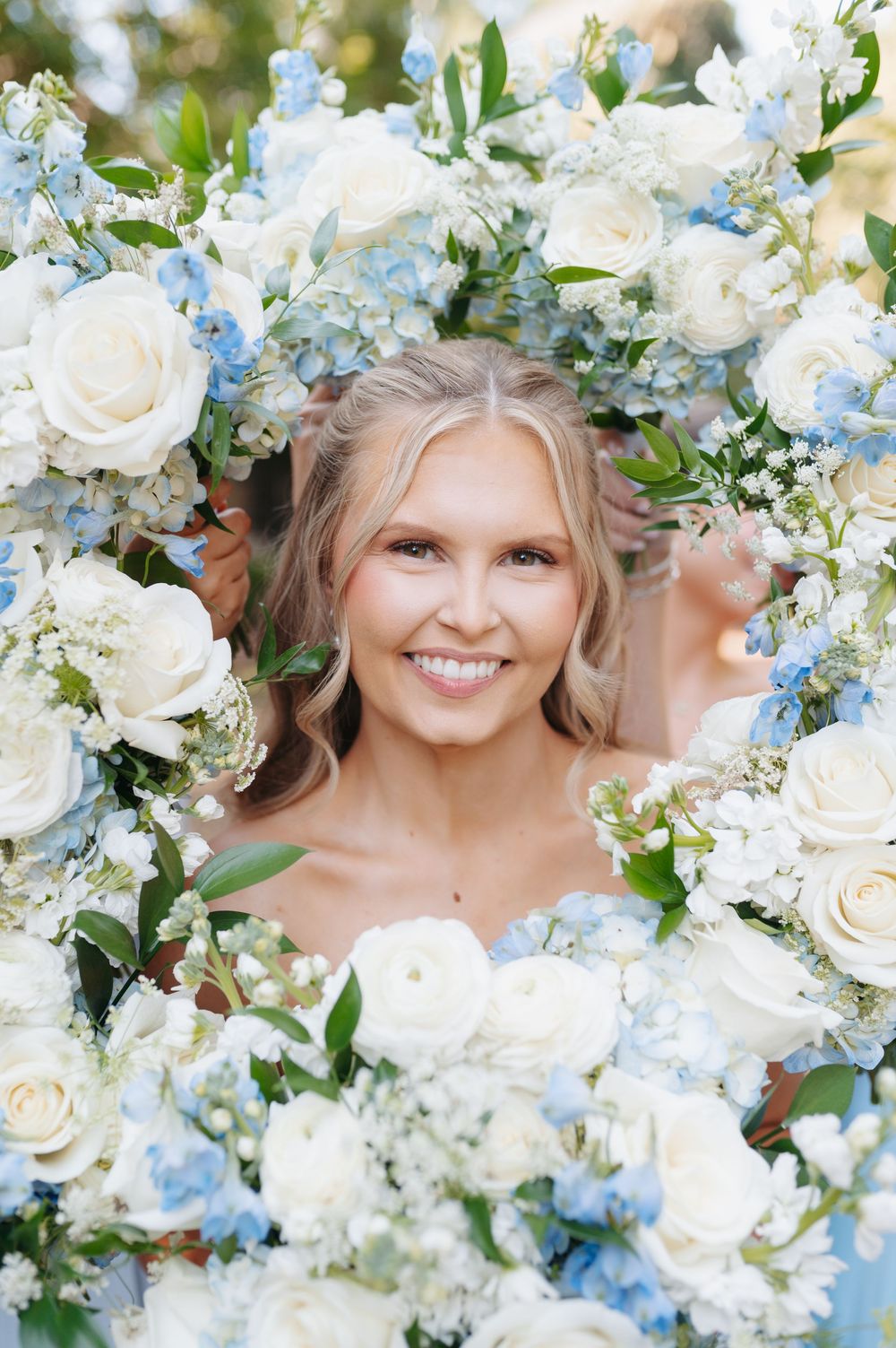 Smiling bride framed by white roses and blue hydrangeas, looking through a floral wreath arrangement.