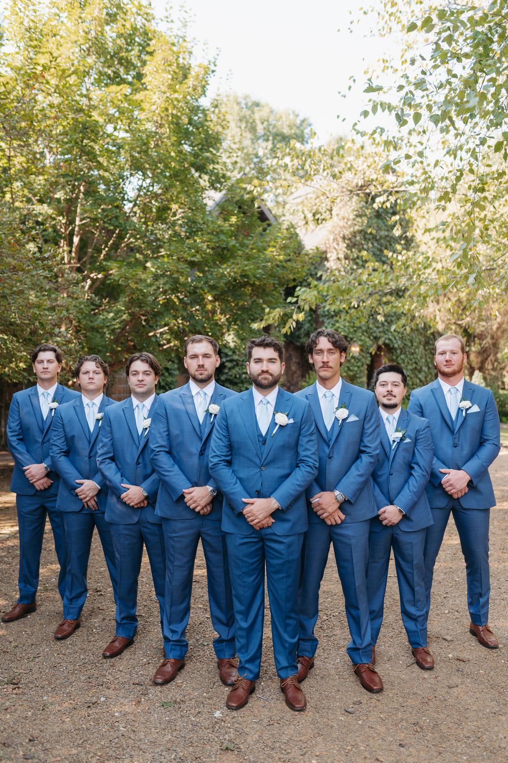 Groom and groomsmen in matching blue suits with white boutonnieres posing outdoors at a wedding.