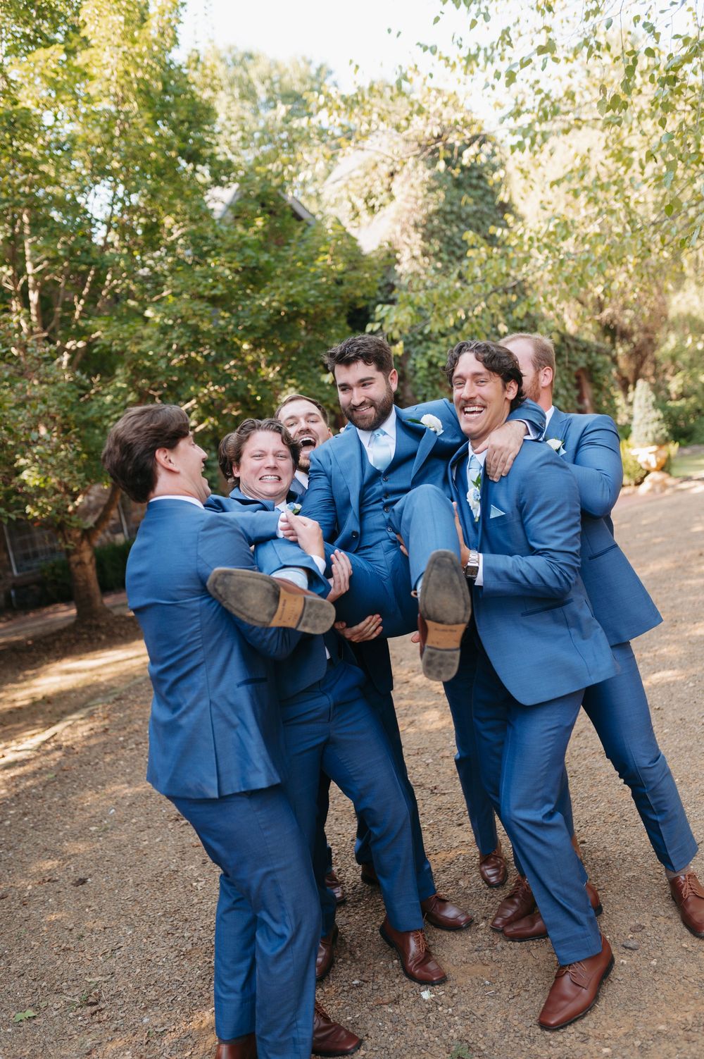 Groomsmen in blue suits joyfully lifting the groom outdoors at a wedding celebration.