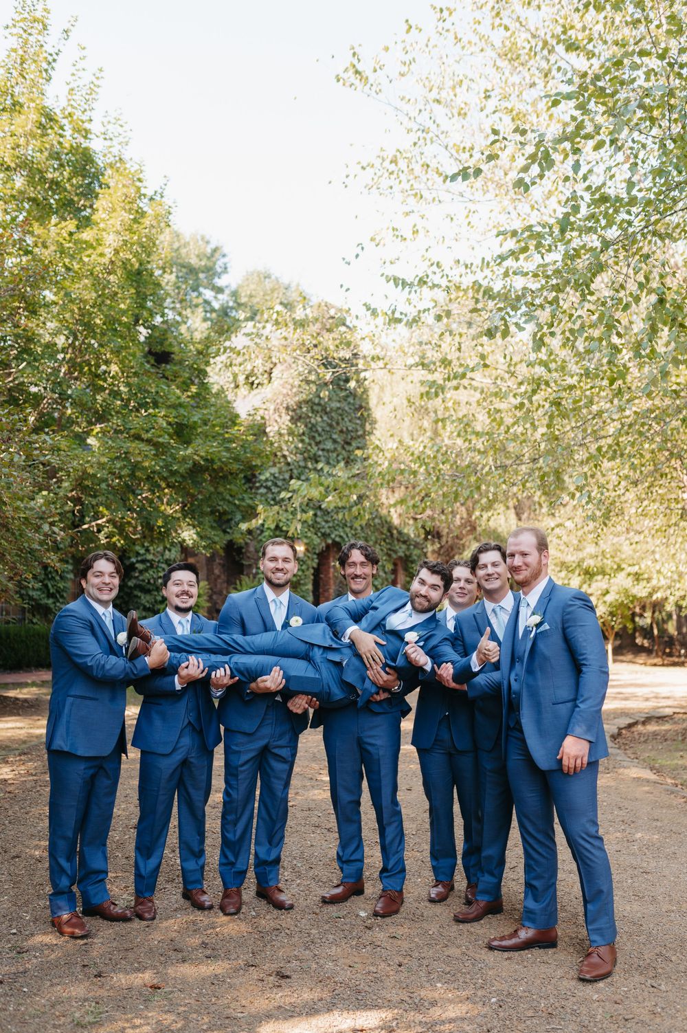 Groomsmen in blue suits lifting the groom outdoors at a wedding, surrounded by lush green trees.