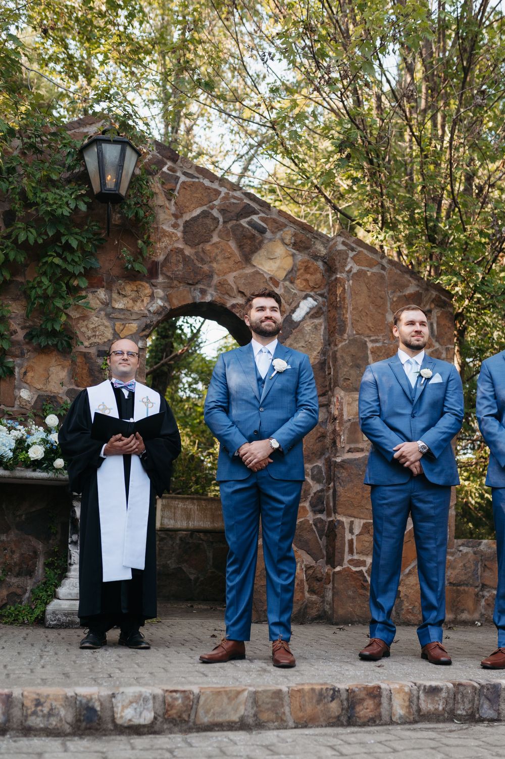 Groom in blue suit stands at altar with groomsmen and officiant at stone arch wedding ceremony.