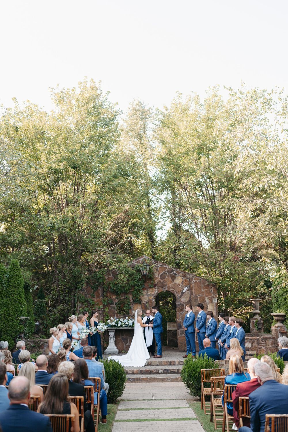 Outdoor garden wedding ceremony with guests seated along an aisle as couple exchanges vows.