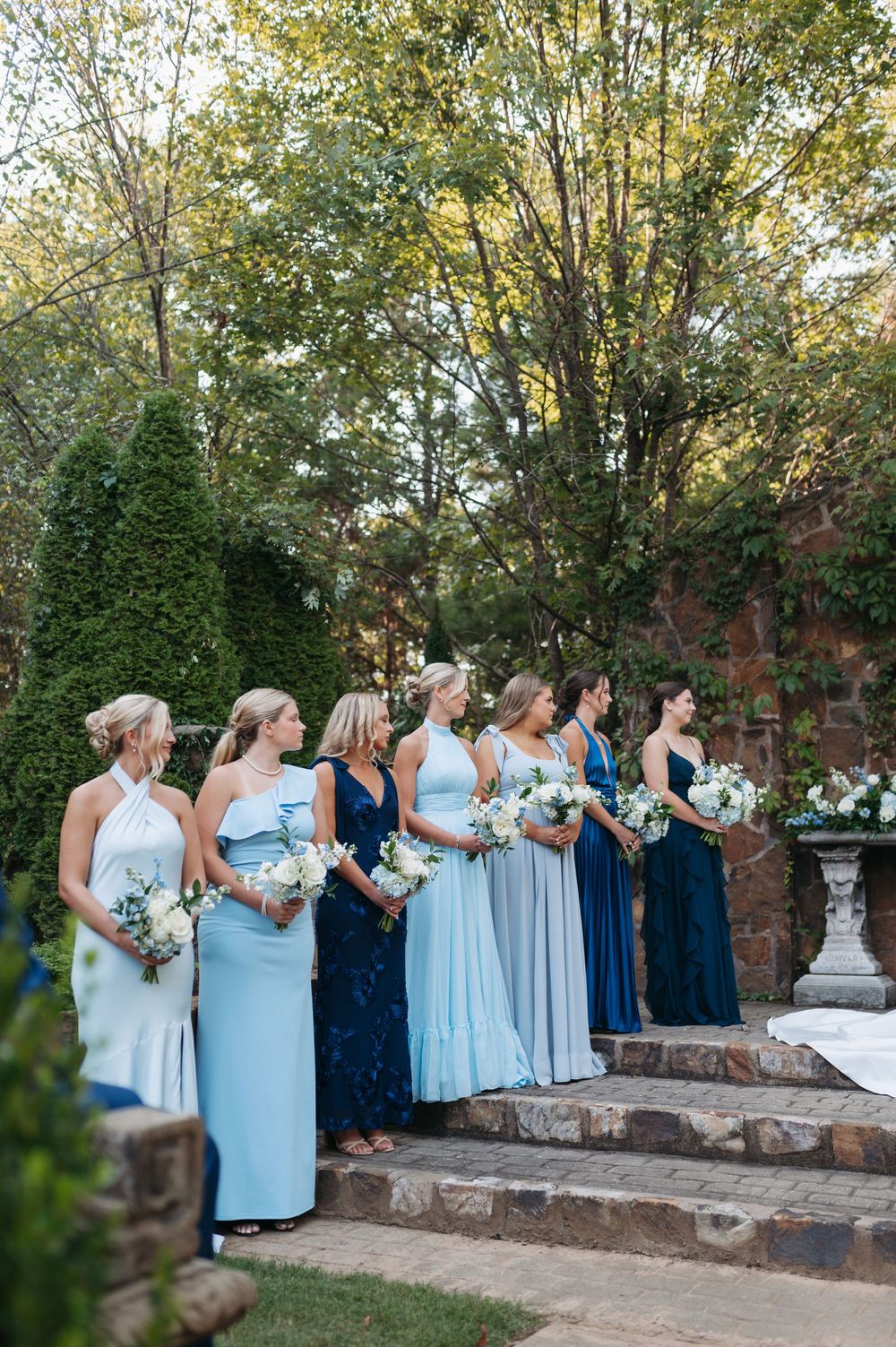 Bridesmaids in mismatched blue gowns holding white bouquets stand on stone steps during outdoor wedding ceremony.