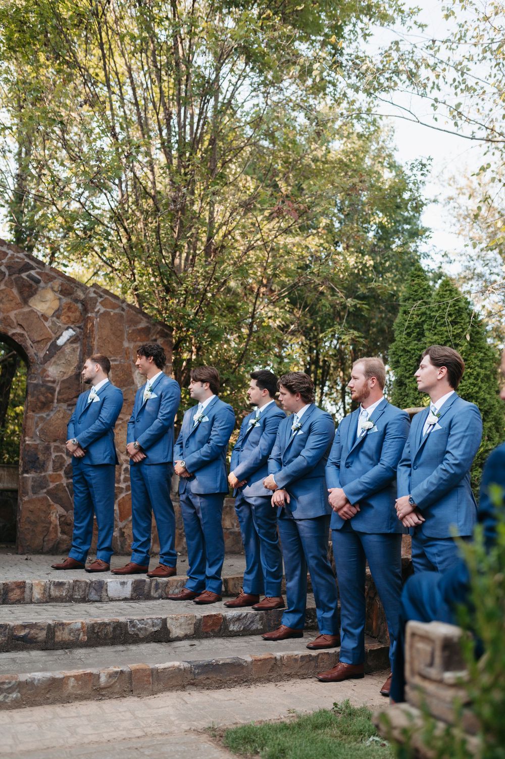 Groomsmen in matching blue suits with white boutonnieres stand at attention on stone steps during wedding ceremony.
