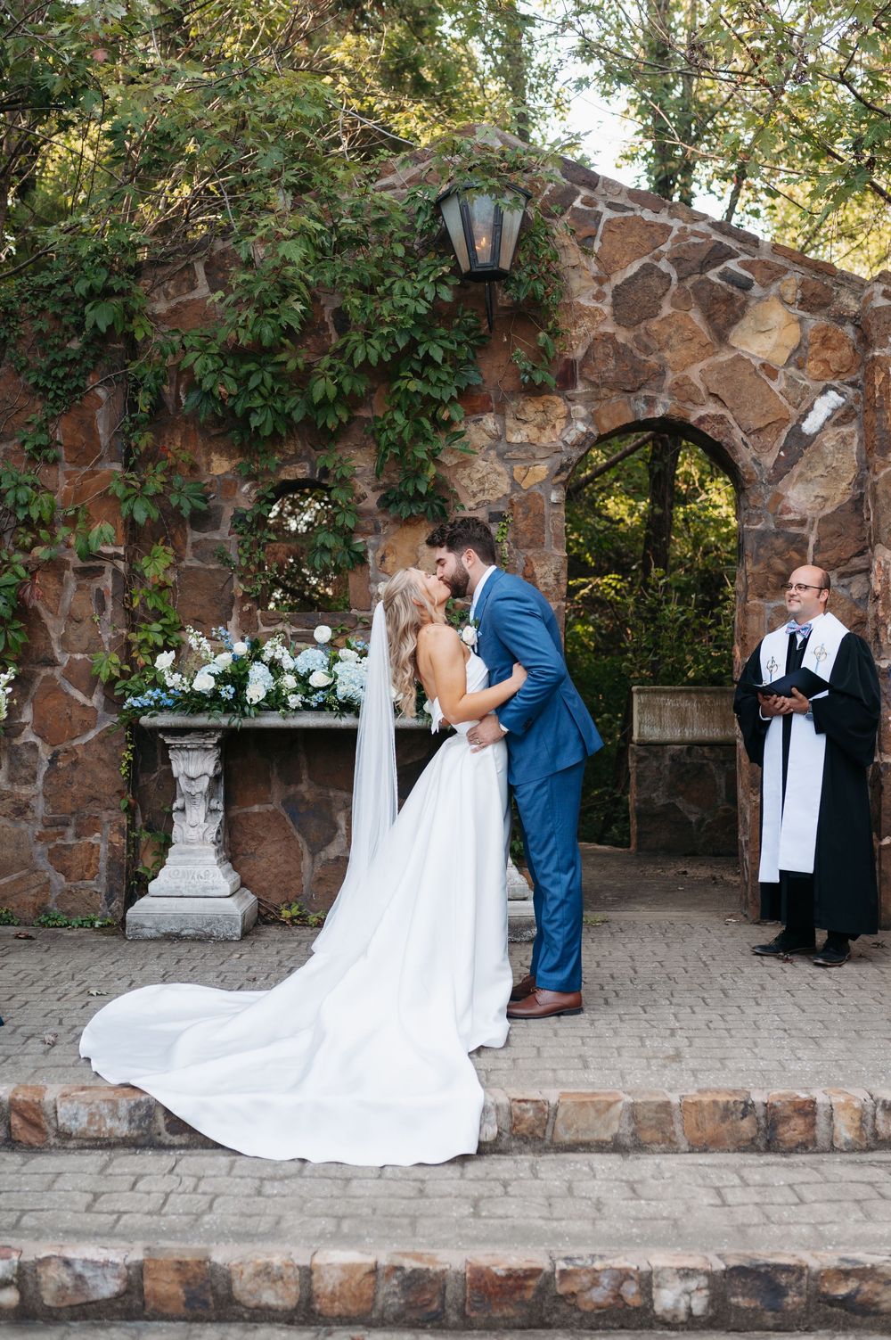 Bride and groom share first kiss at outdoor stone arch wedding ceremony while officiant watches nearby.