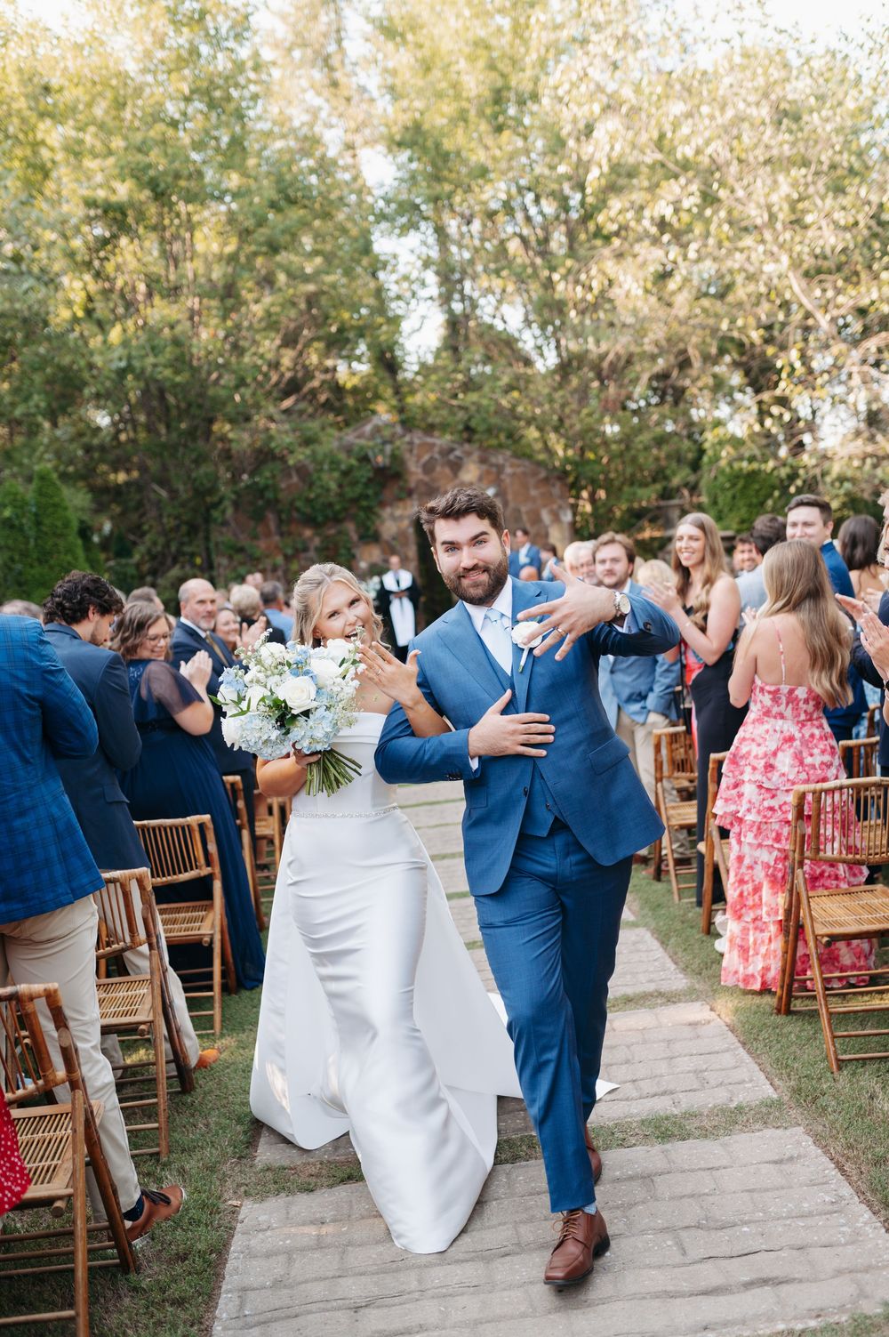 Bride and groom walk down outdoor wedding aisle, smiling at guests seated in wooden chairs amid lush greenery.