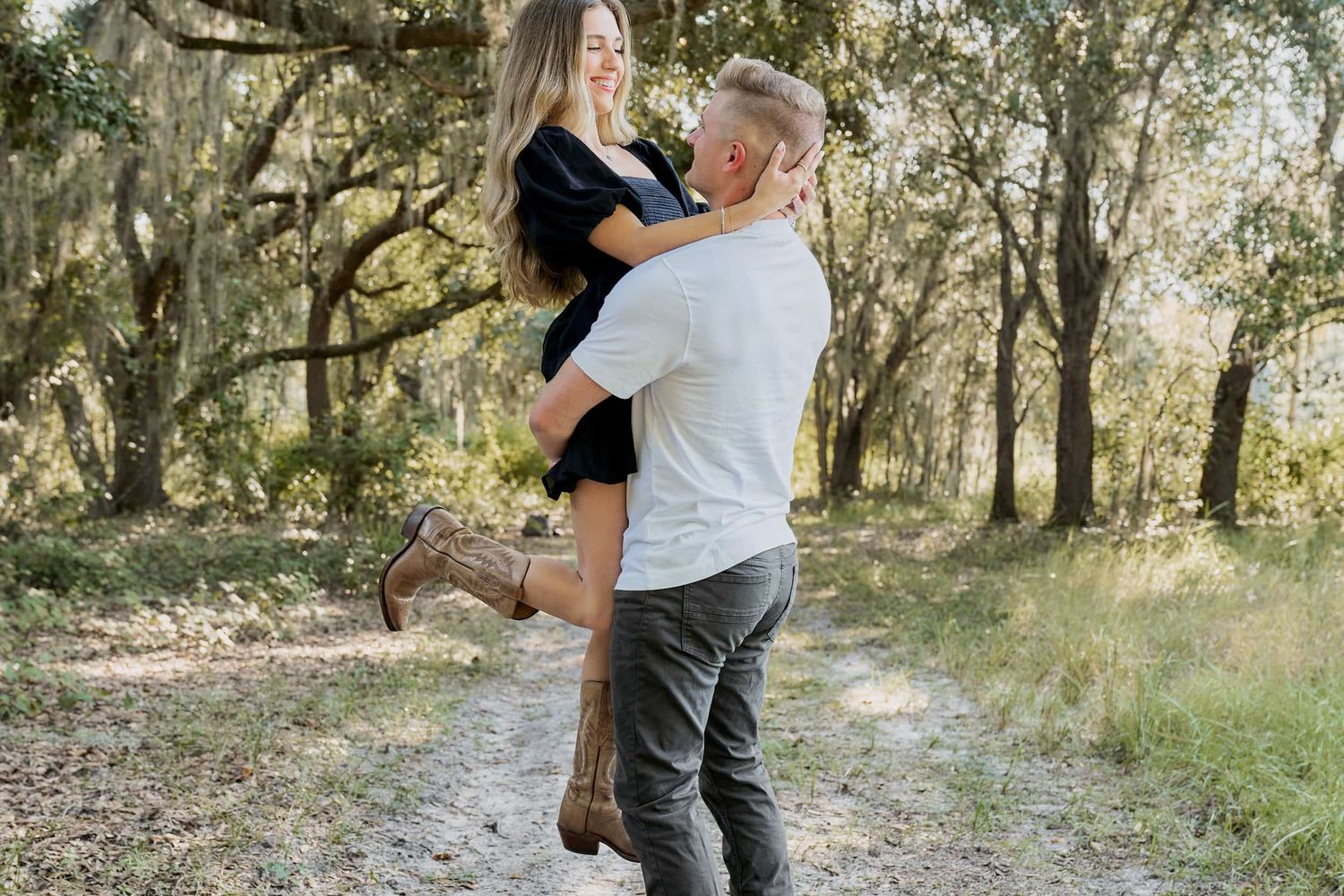 Couple embracing outdoors on a forest path, man lifting woman in a romantic pose among moss-covered trees.