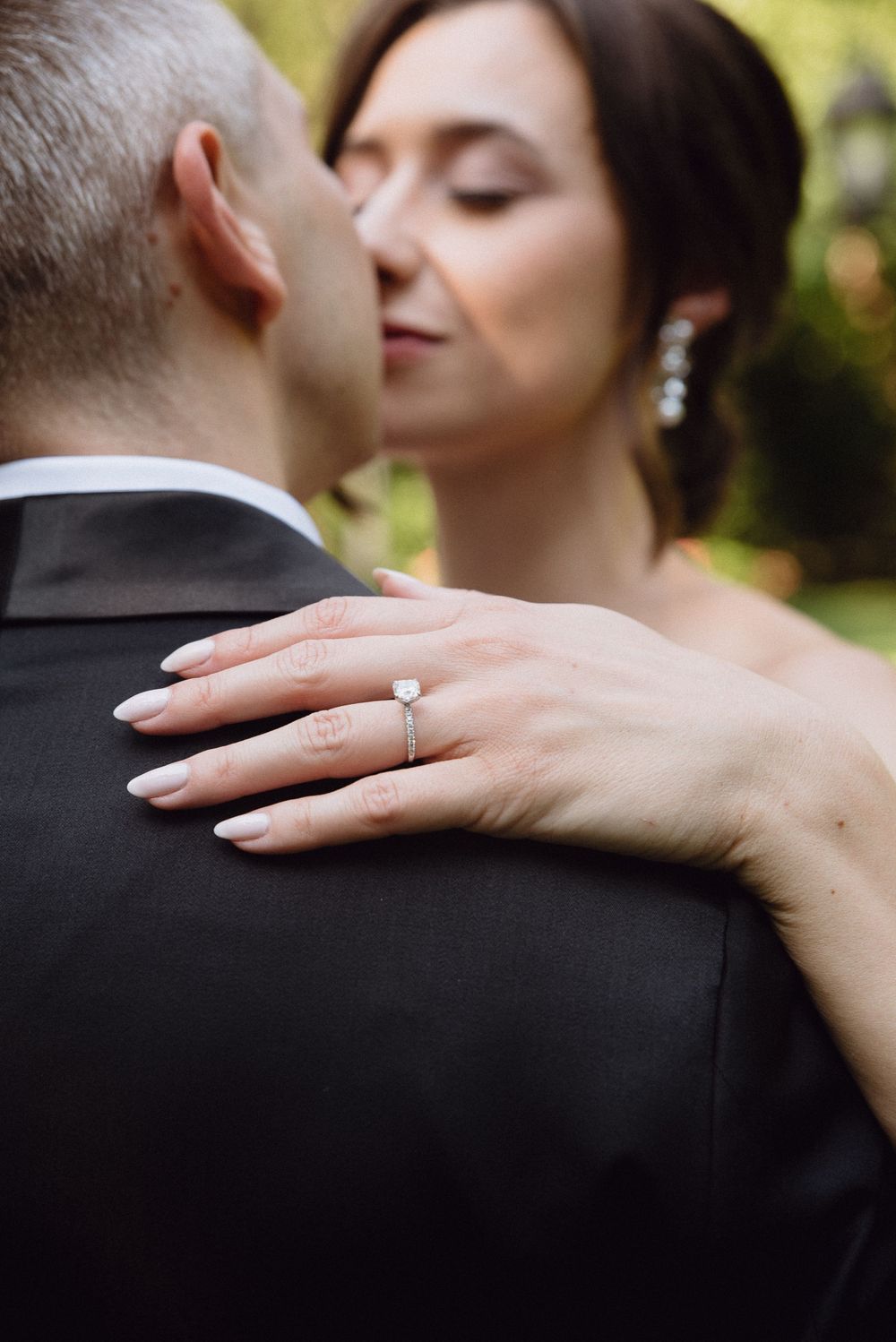 A close-up shot showing a delicate engagement ring and manicured hand resting on a black tuxedo jacket.