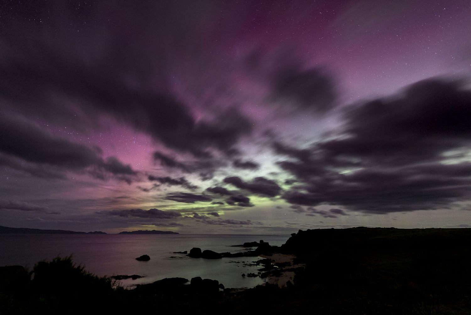Aurora borealis illuminates a dramatic purple and pink sky over a rocky coastline with dark clouds and calm ocean waters.