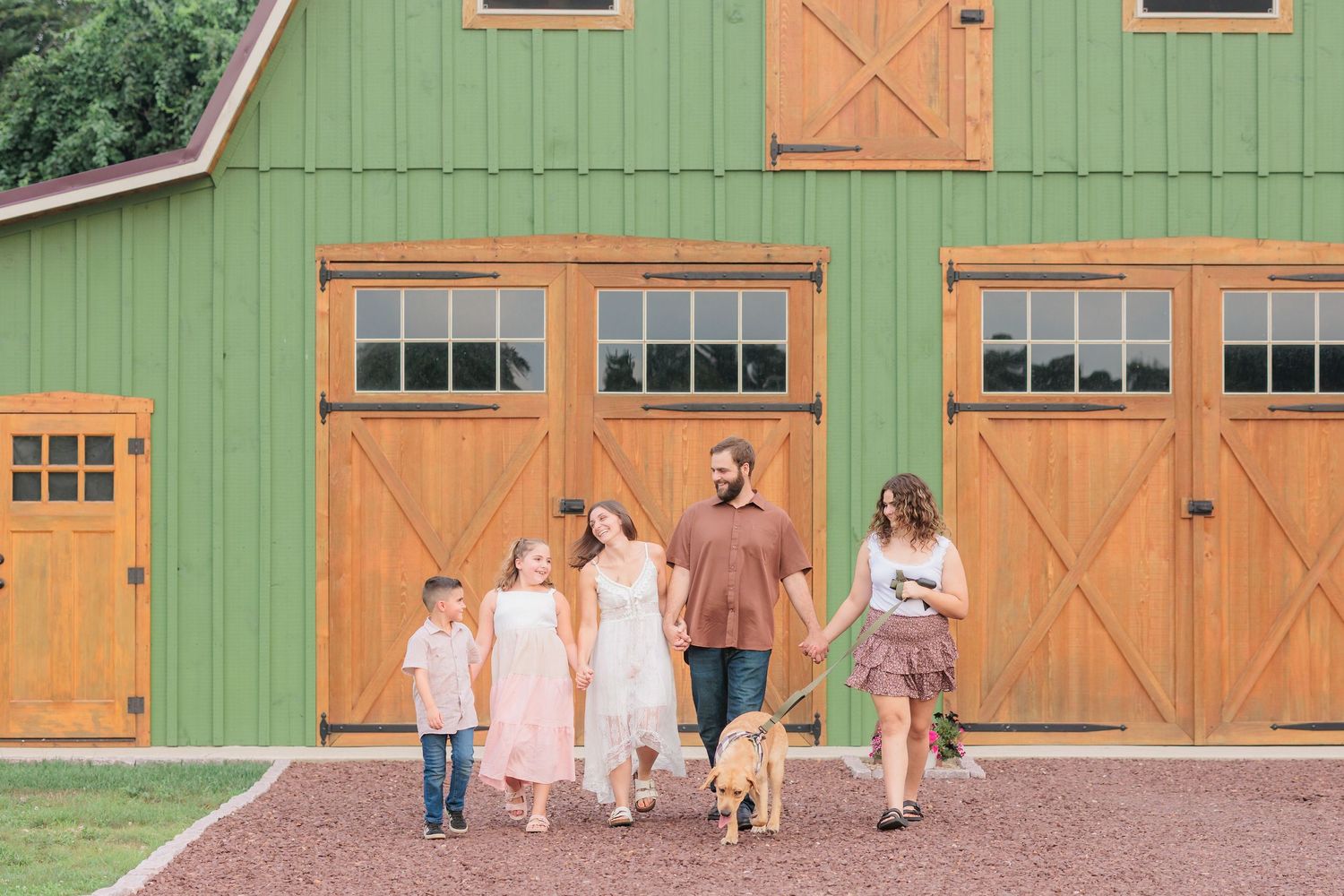 Family of four with dog walking in front of a green barn with wooden doors on a sunny day.