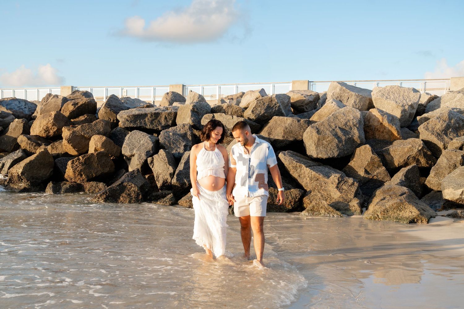 Couple holding hands walking in shallow ocean waves near rocky jetty on sunny beach day.