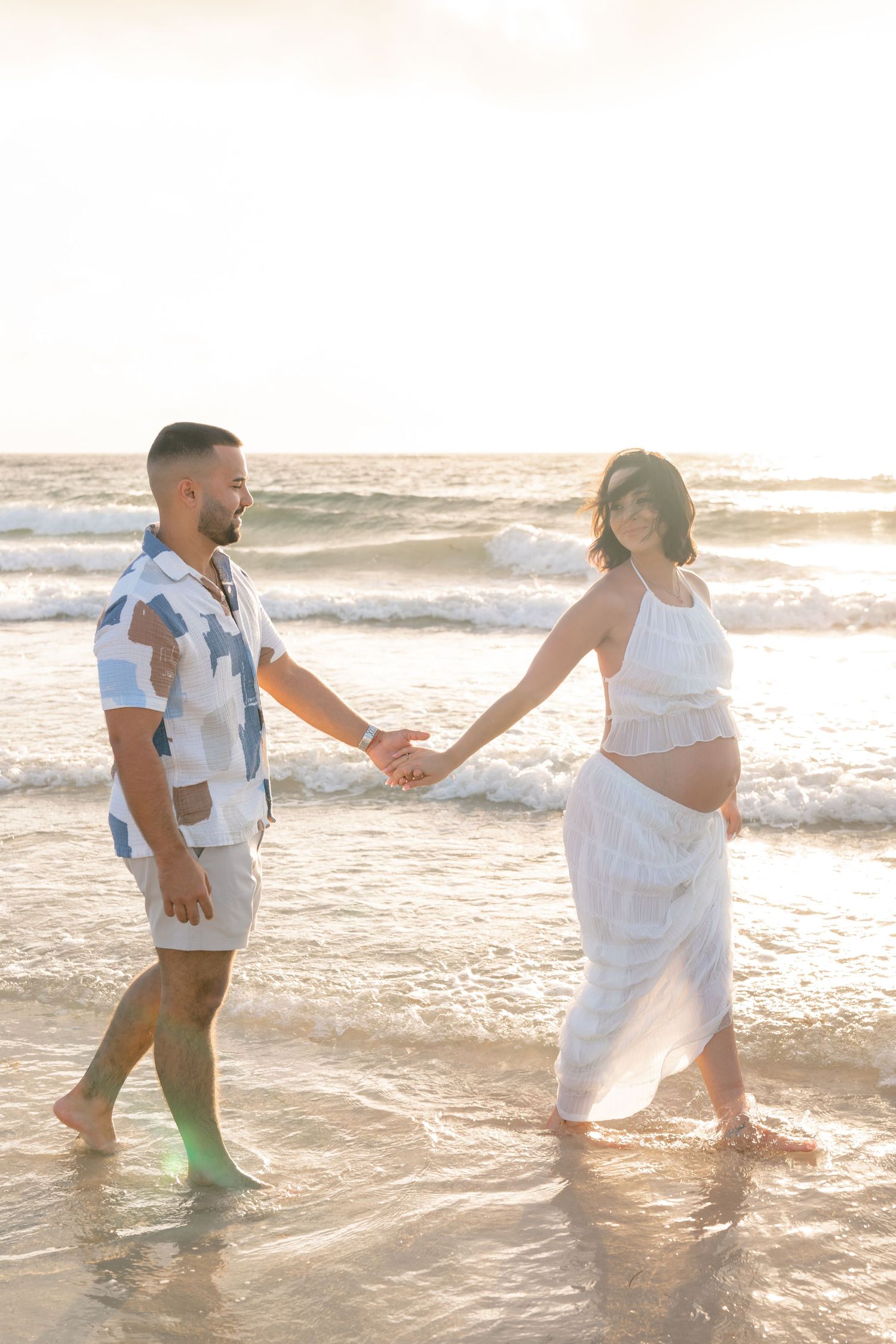 Pregnant couple holding hands walking on a sunlit beach, woman in white dress cradling baby bump, man in patterned shirt.