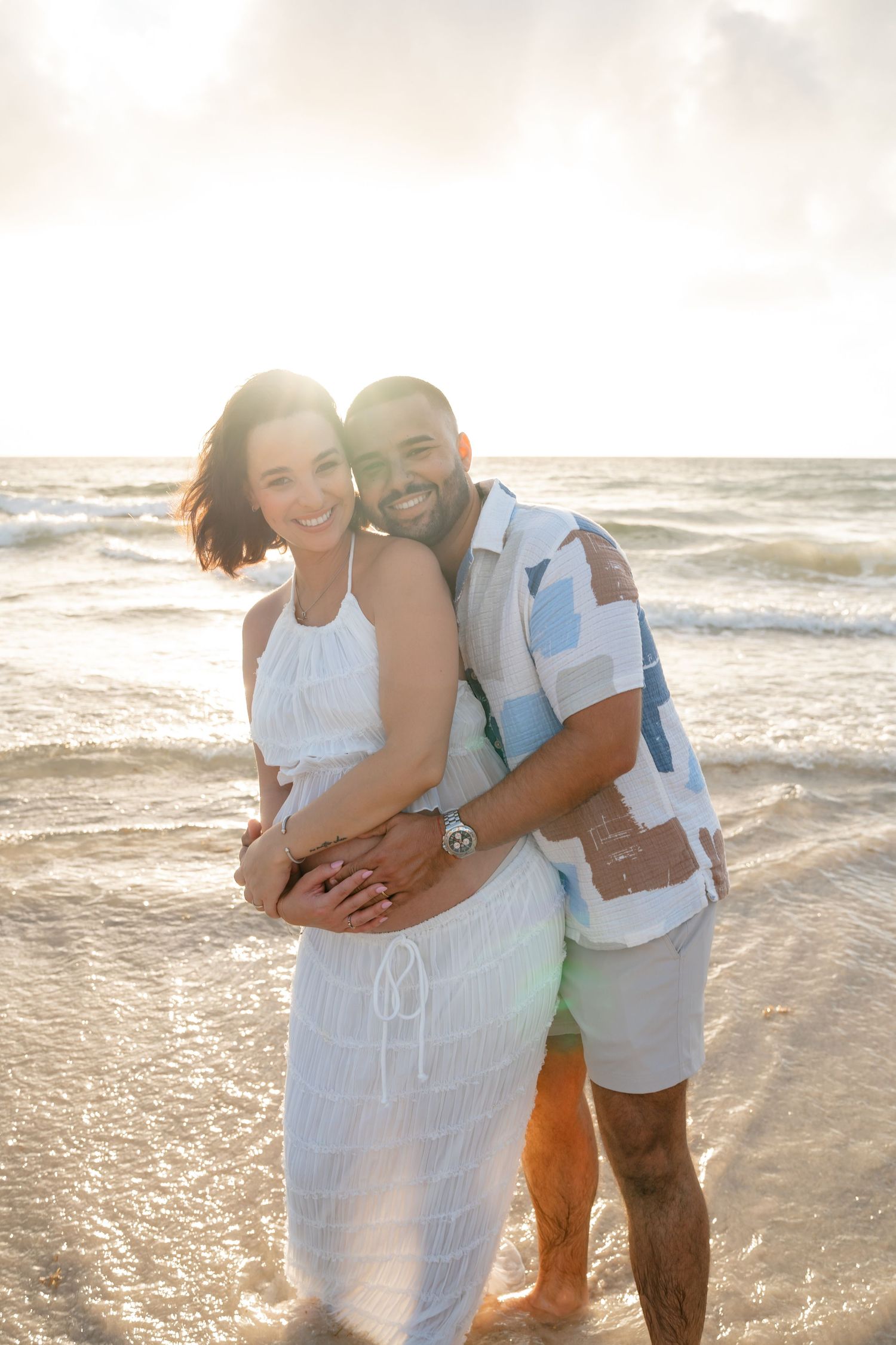 Happy couple embracing on a sunny beach, woman in white dress and man in colorful patterned shirt smiling together.