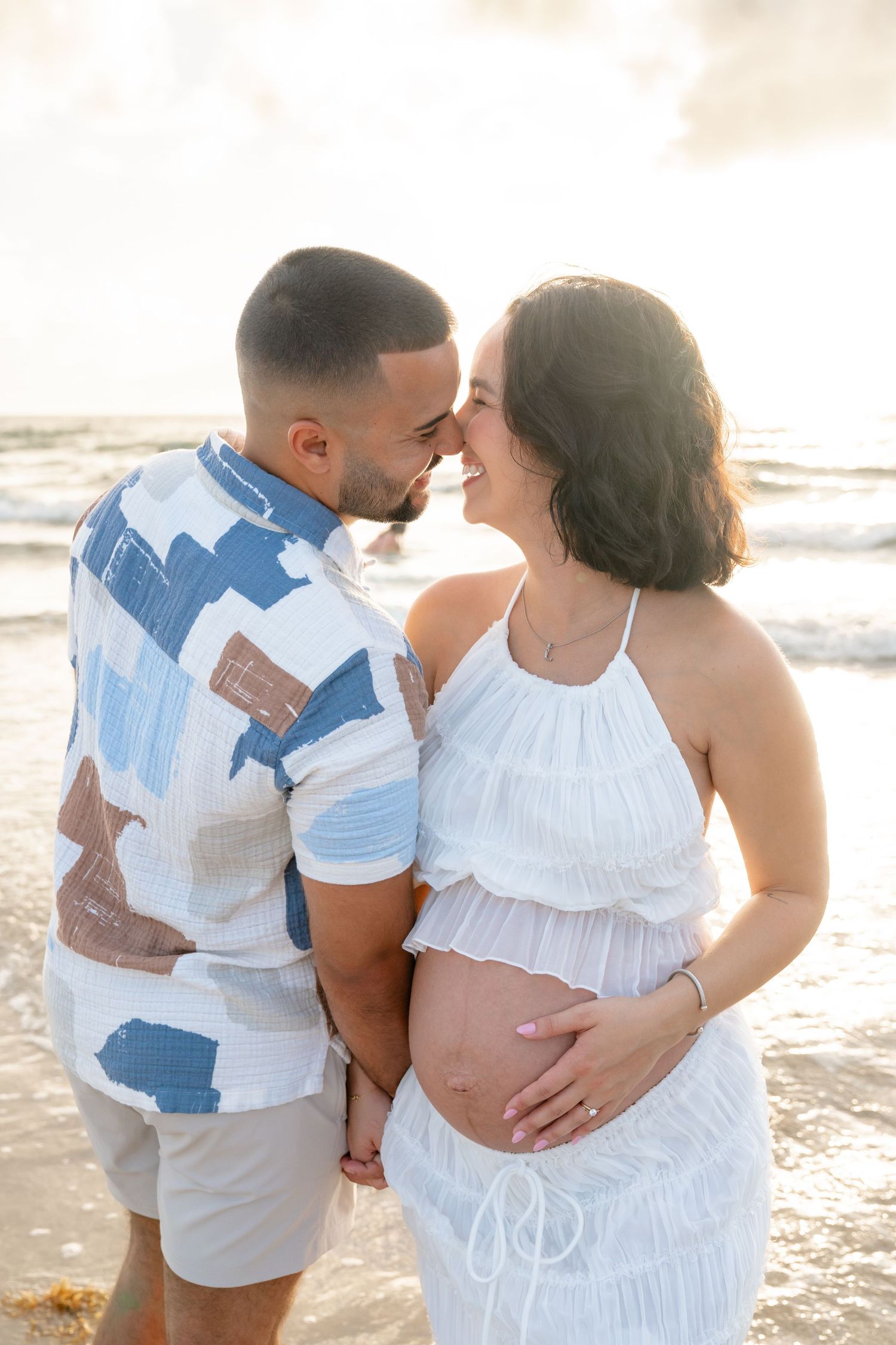 Couple sharing a tender moment on the beach at sunset, with pregnant woman in white dress cradling her baby bump.