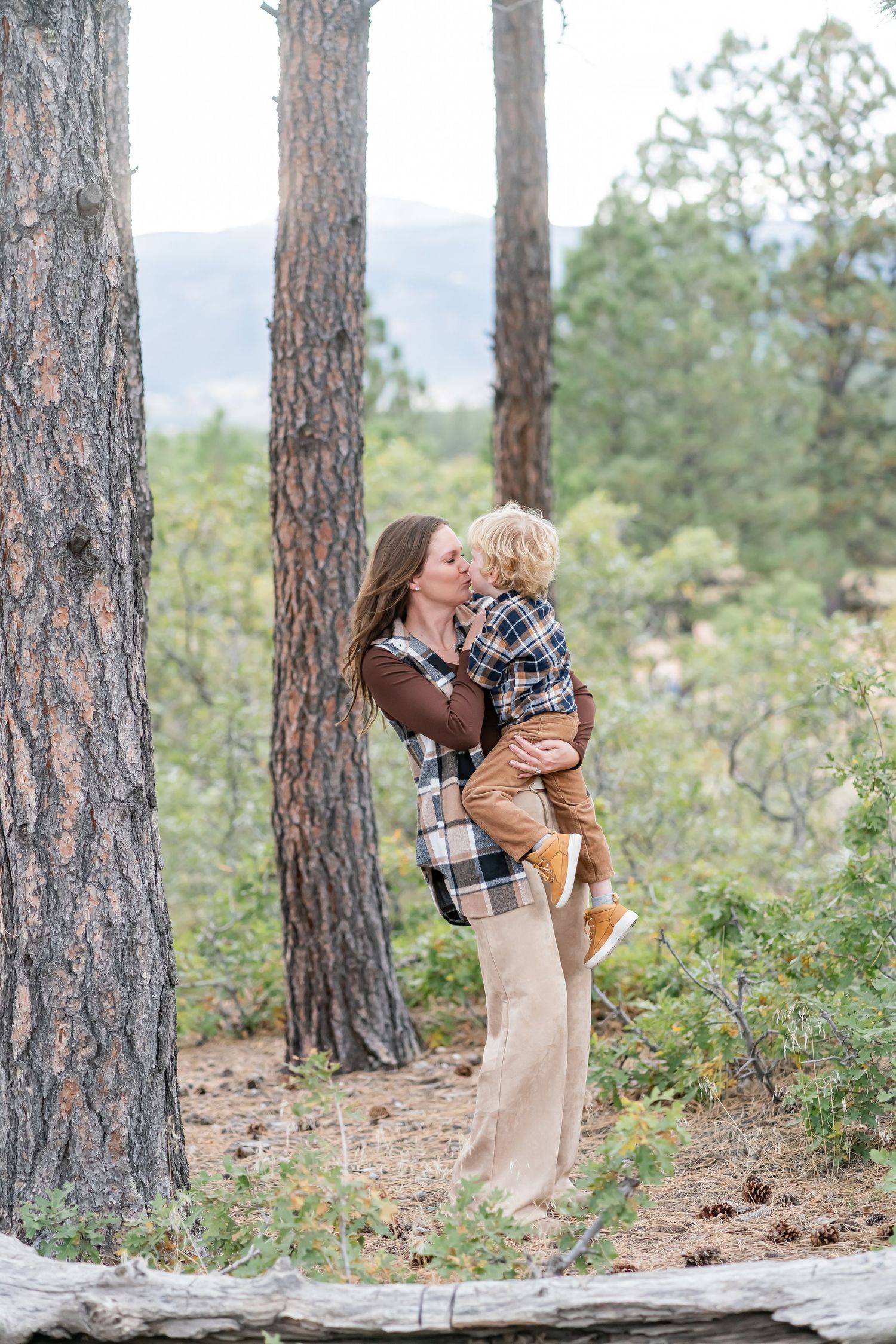 A mother lovingly kisses her toddler son while holding him in a scenic forest setting.