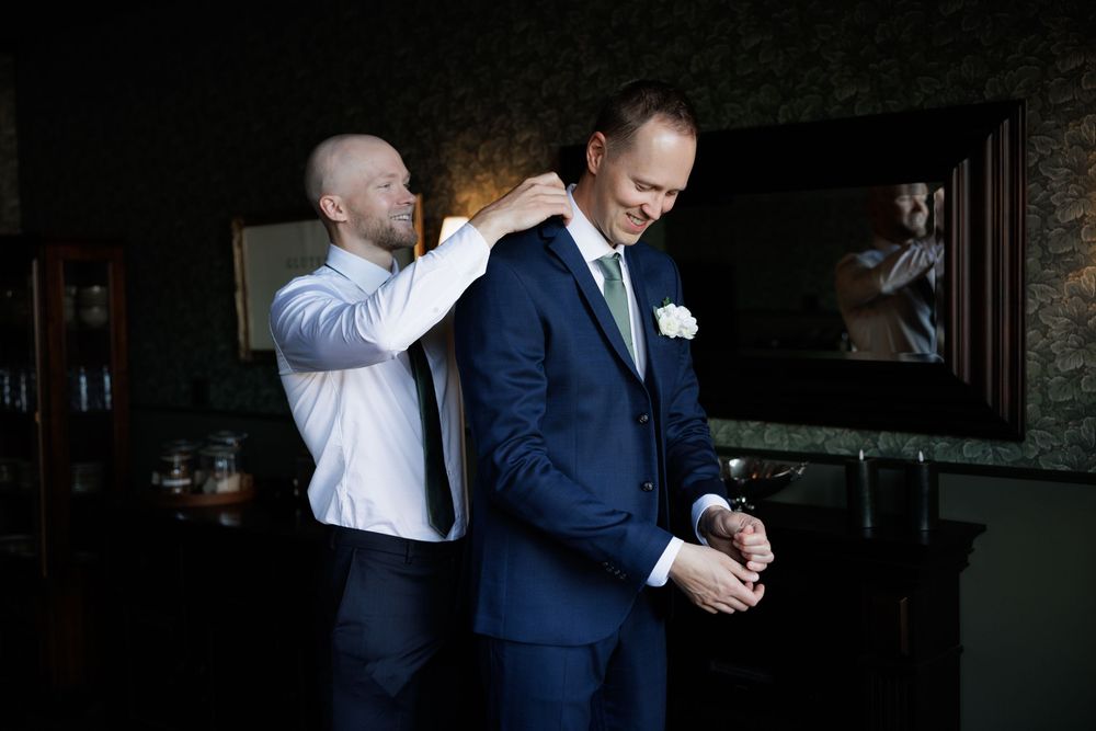A groomsman adjusts the collar of a groom in a navy suit before a wedding at Jonsereds Fabriker and getting ready at Gibssons Hotel