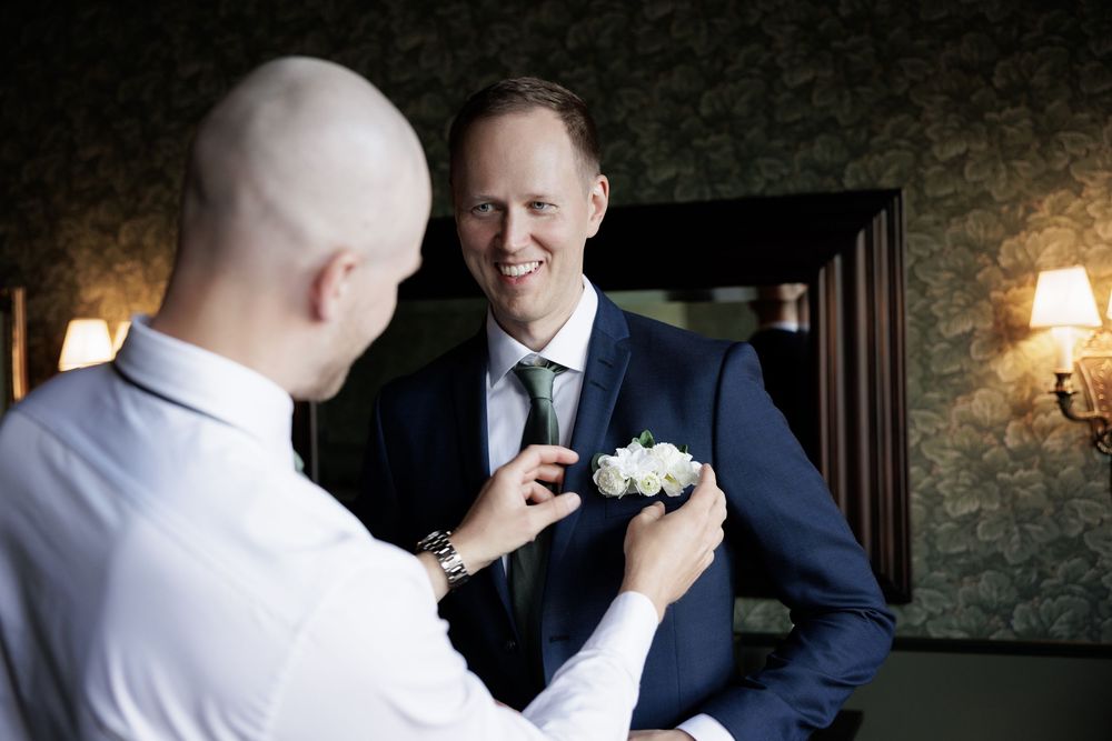 Groom smiling as best man pins white boutonniere on navy suit lapel in elegant room with wallpaper and warm lighting.