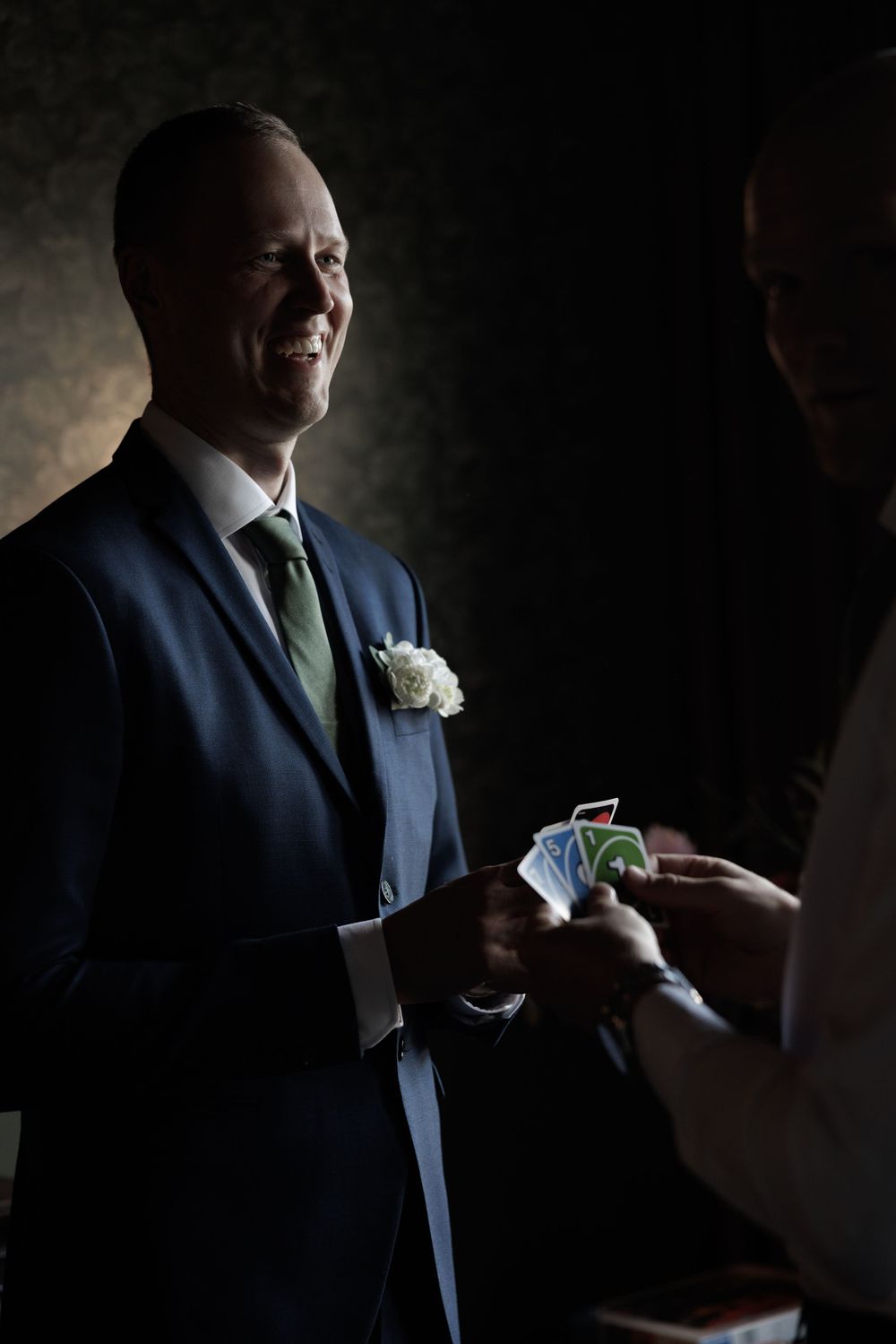 Smiling groom in navy suit with white boutonniere receiving playing cards during wedding celebration.