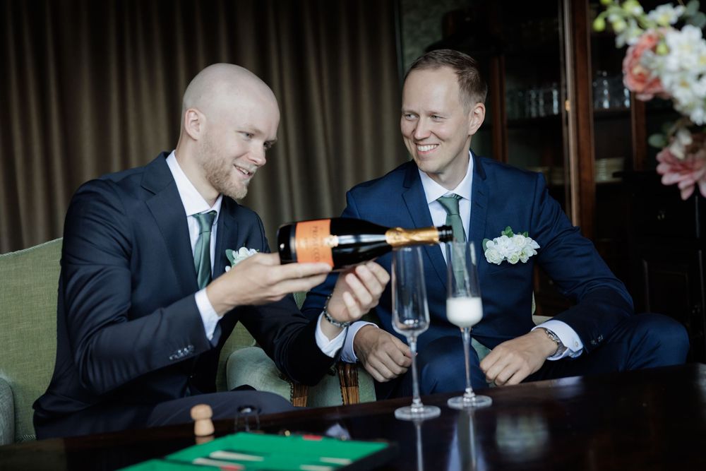 Two grooms in navy suits share a champagne toast at their wedding reception at Gibssons hotel Jonsereds Fabriker