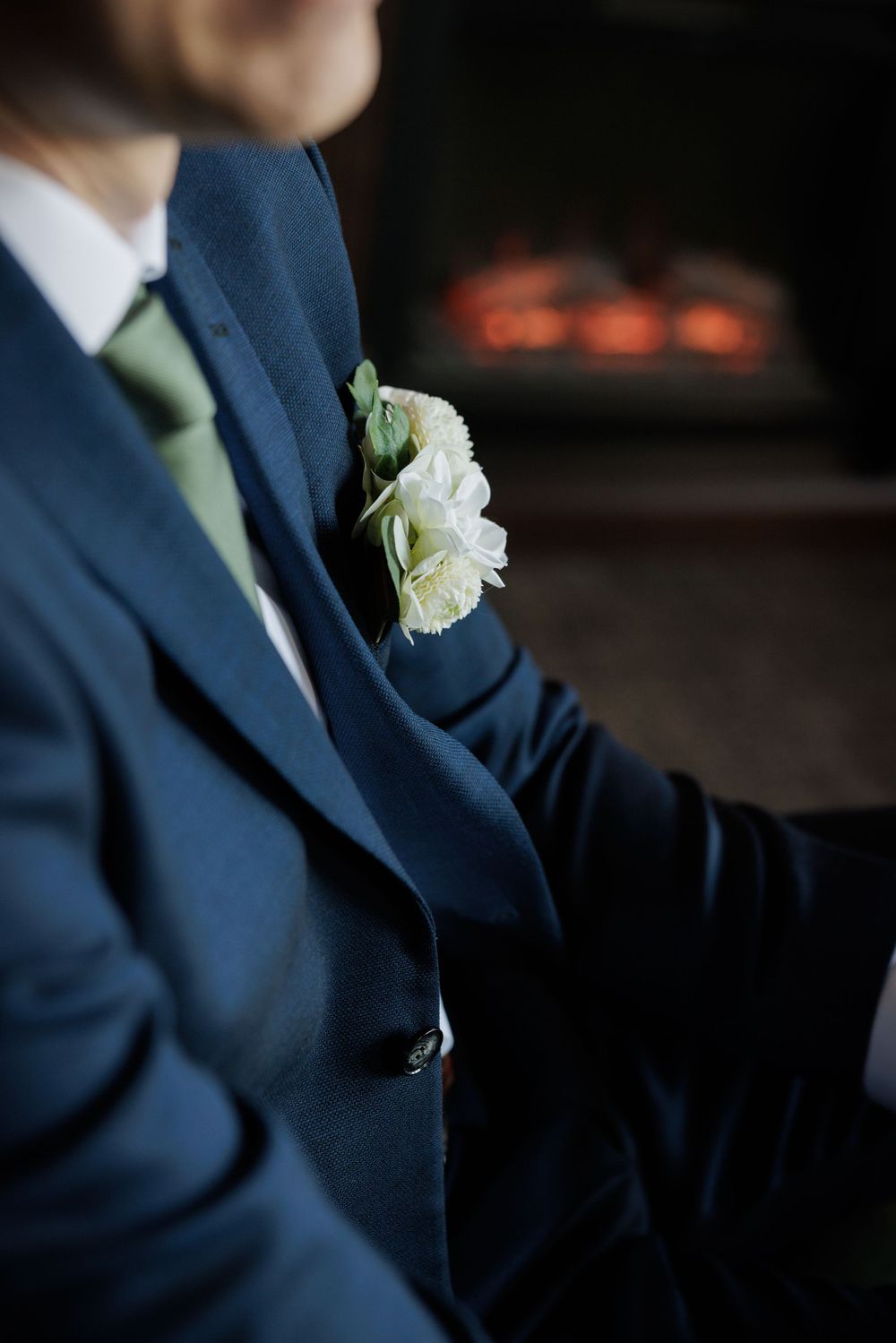 Groom in navy suit with green tie and white carnation boutonniere, seated near a glowing fireplace.