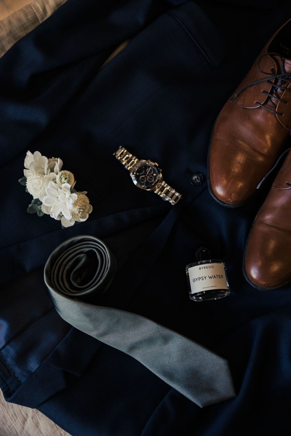 Groom wedding flat lay with navy suit, gold watch, gray tie, boutonniere, brown leather shoes, and Byredo Gypsy Water cologne.