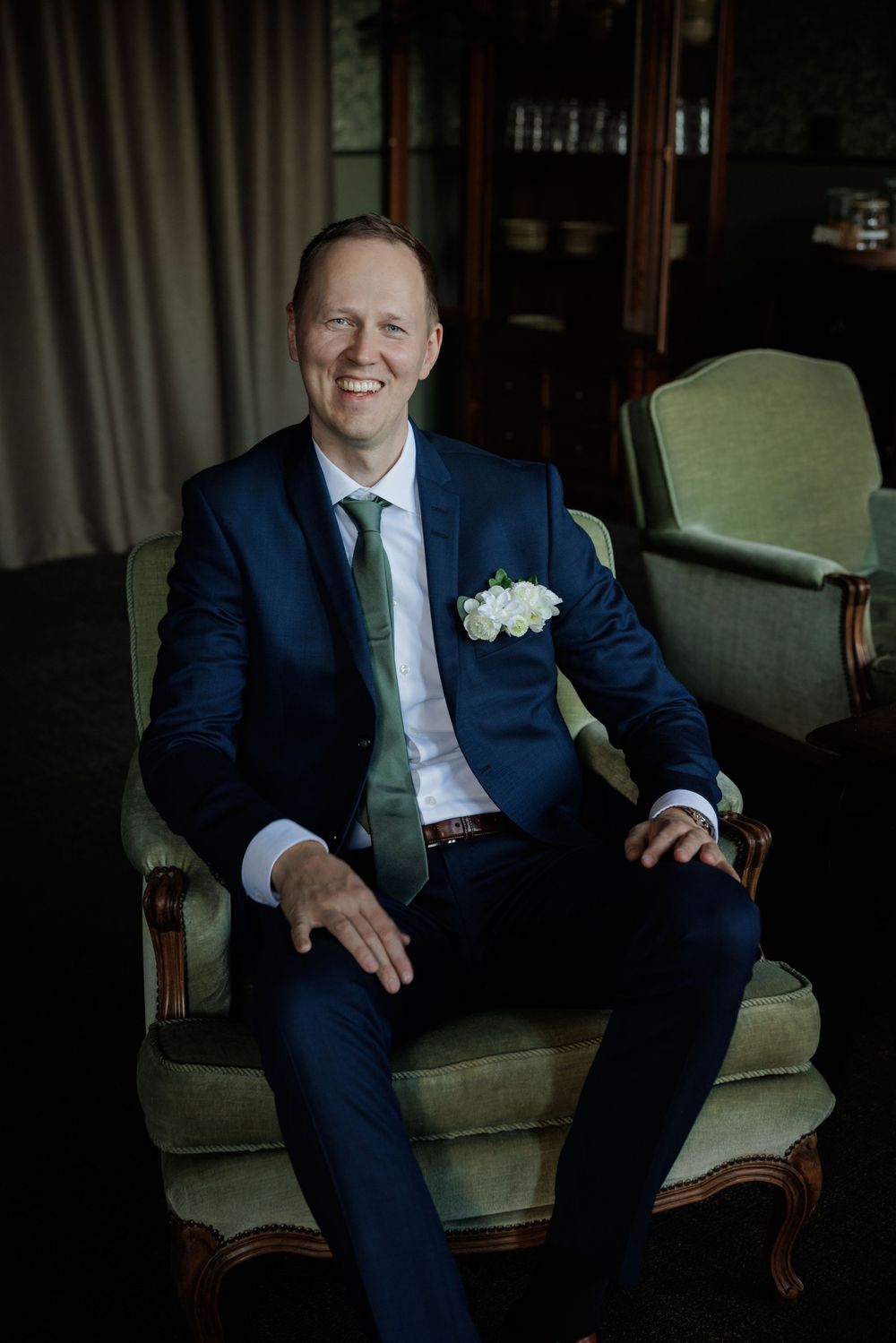 Smiling groom in navy suit with white boutonniere seated in green armchair at wedding venue.