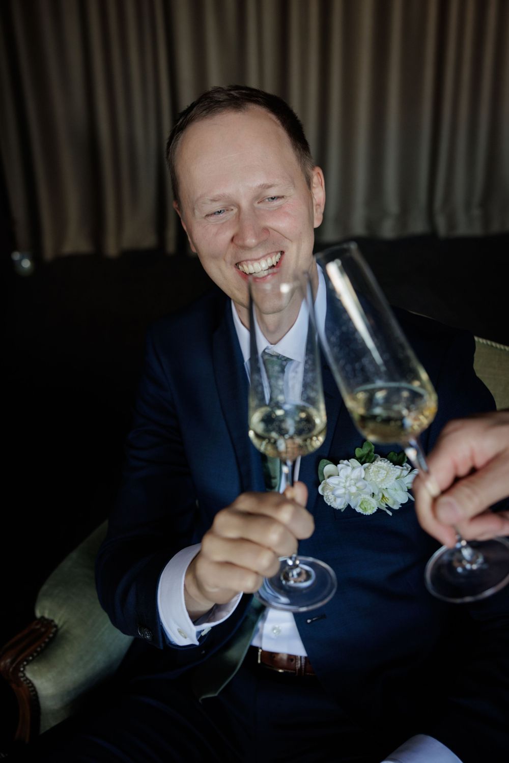 Smiling groom in navy suit toasting with champagne flutes, wearing floral boutonniere at wedding reception.