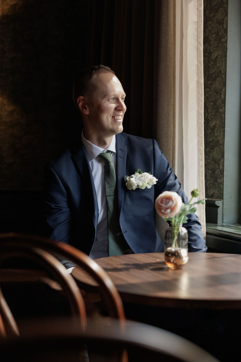 Smiling groom in navy suit with white boutonniere sits by window at wooden table with flower vase.