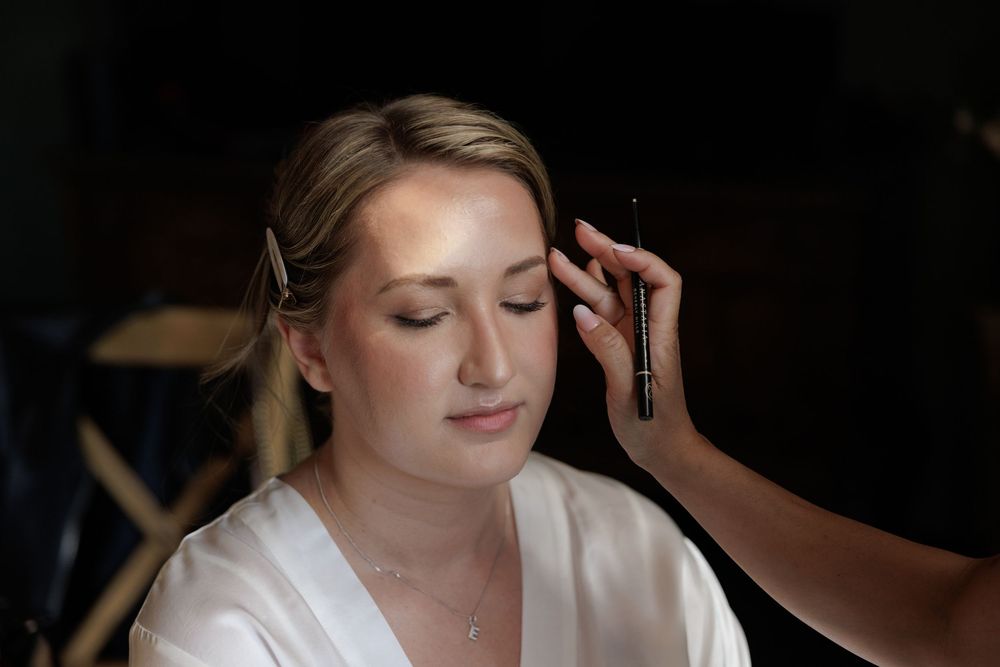 Bride getting eyebrows done by makeup artist while wearing white satin robe during wedding day preparation.