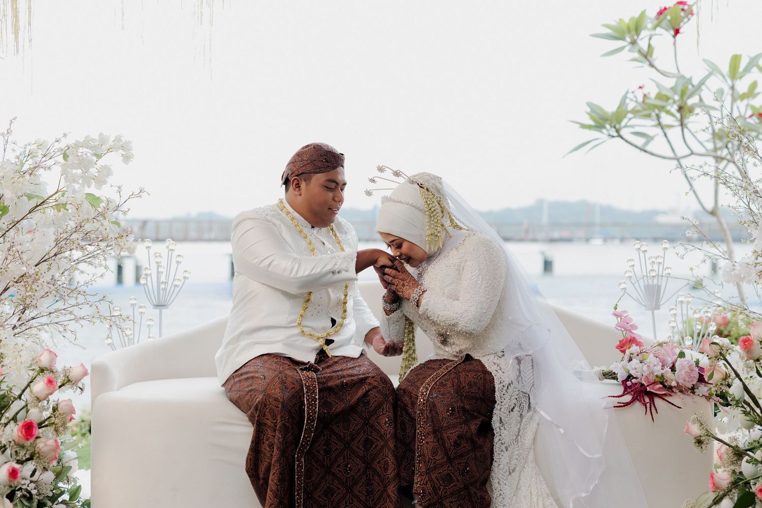 Muslim couple in traditional Javanese wedding attire sitting together at an outdoor ceremony with floral decorations and ocean view.