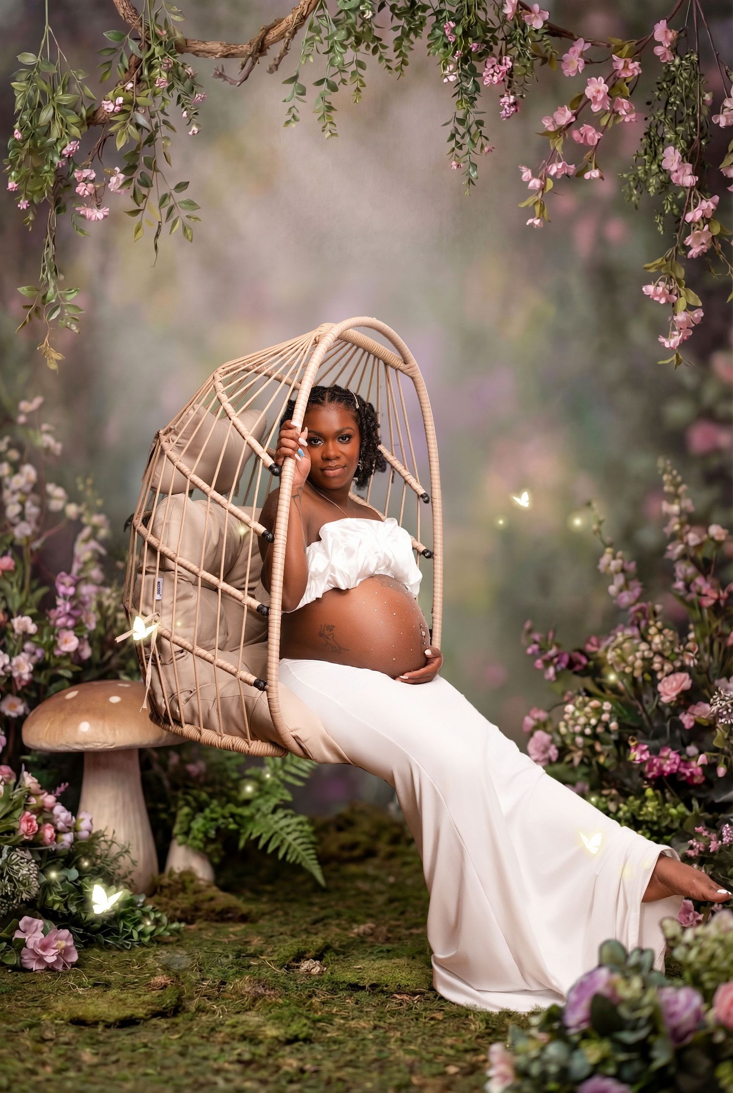 Pregnant woman in white dress sitting in rattan swing chair surrounded by flowers and mushrooms in enchanted forest setting.