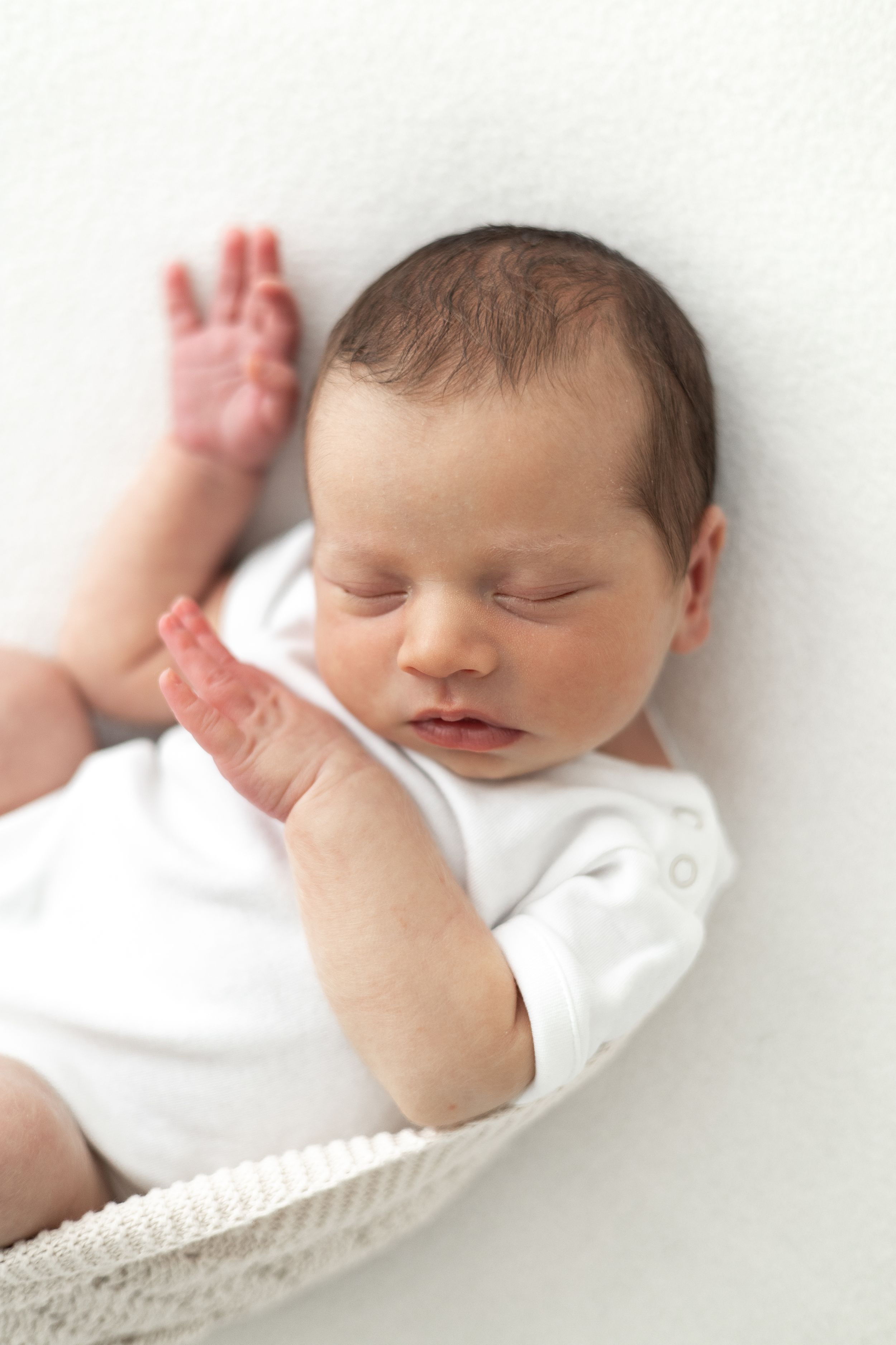 Peaceful newborn baby sleeping in a white blanket, wearing a white onesie, with tiny hands raised newborn photoshoot Nottingham