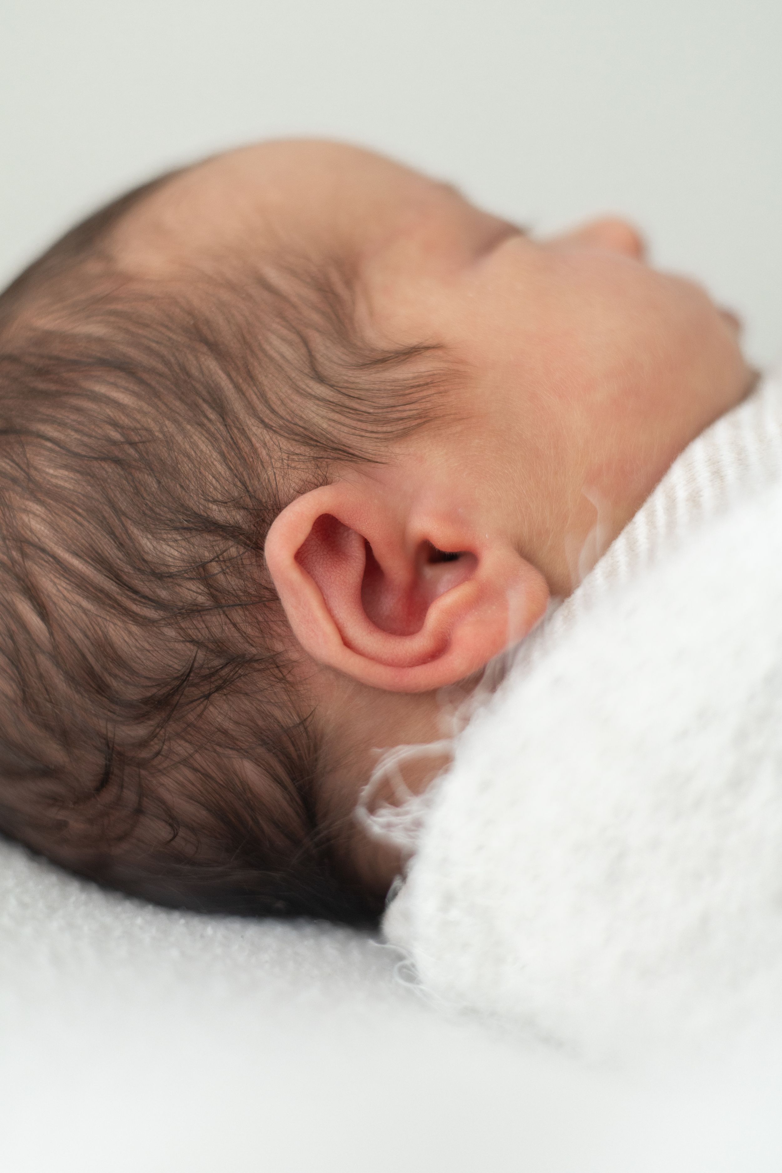 Close-up of a sleeping newborn baby with dark hair, showing ear detail, wrapped in a soft white blanket newborn photoshoot Nottingham