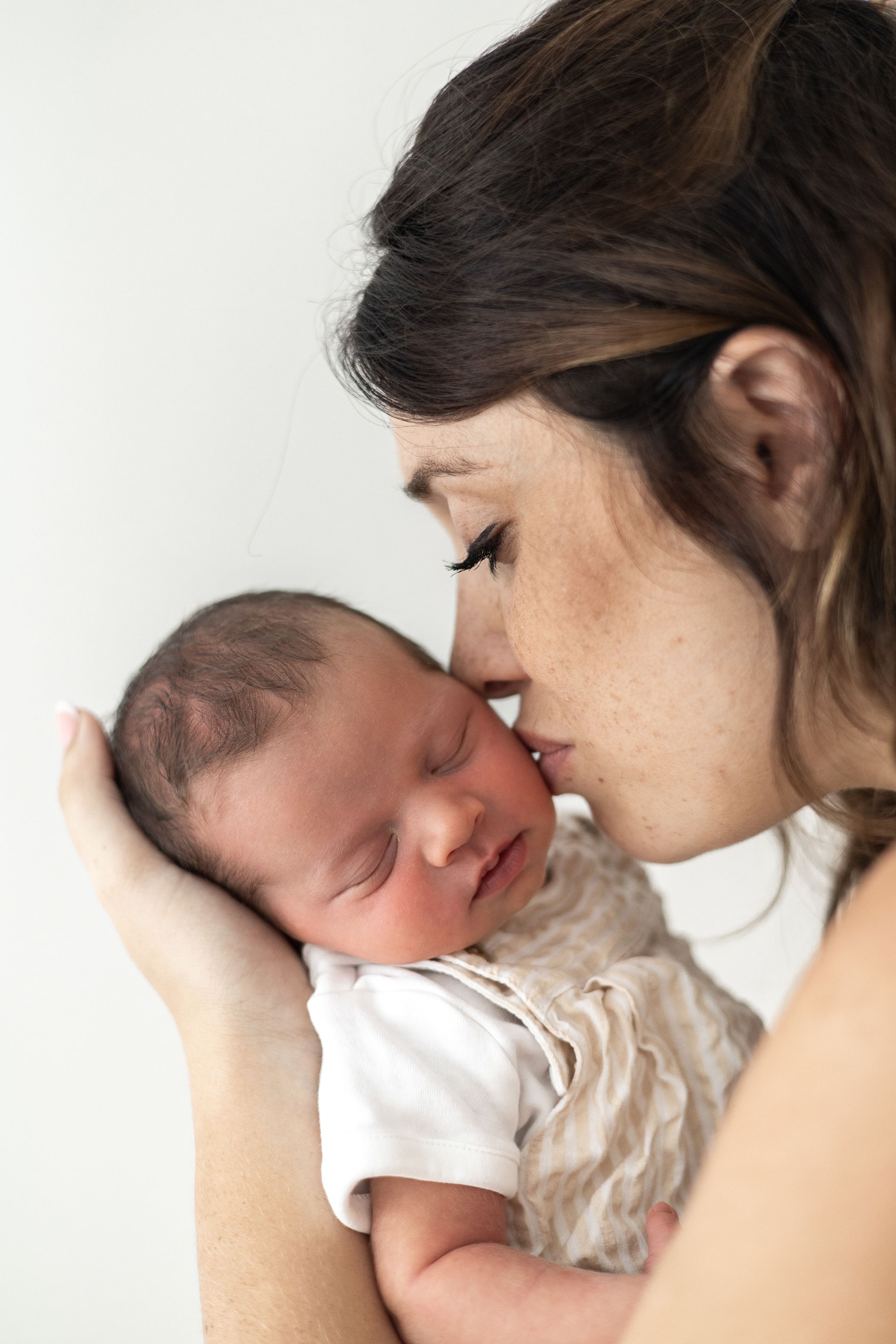 A mother gently kisses her sleeping newborn baby, cradling the infant tenderly in her hands newborn photoshoot Nottingham