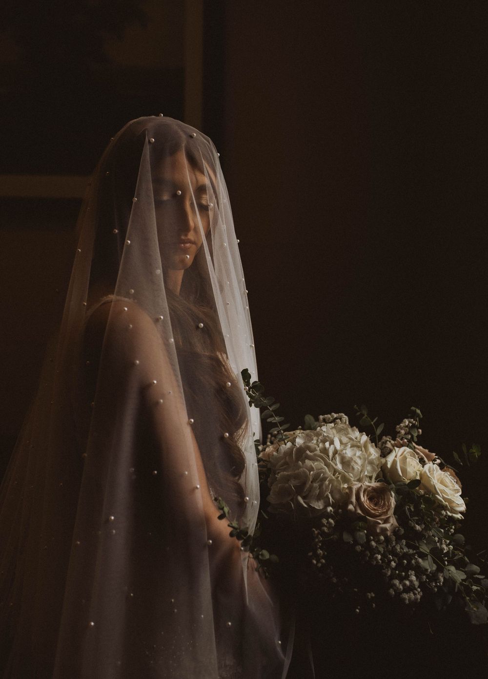 Bride wearing a pearl-embellished wedding veil holding a bouquet of white and pink roses in dramatic lighting.