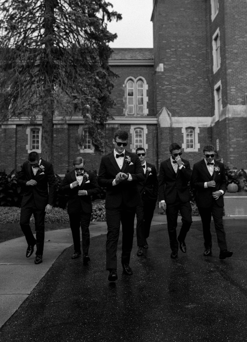 Groom and groomsmen in black tuxedos walking confidently outside a brick building in a black and white wedding photo.