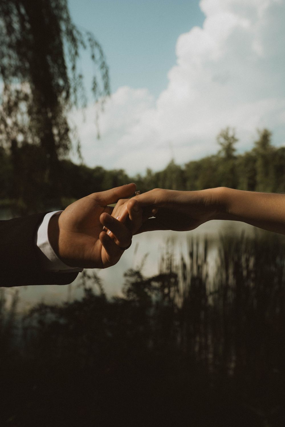 Two hands gently touching fingertips outdoors near a lake with willow trees and cloudy sky in background.