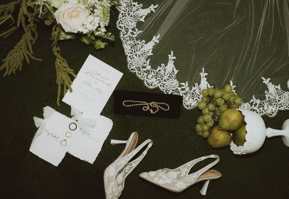 Elegant wedding flat lay with lace veil, bridal heels, rings, invitations, necklace, and fresh grapes on dark green background.