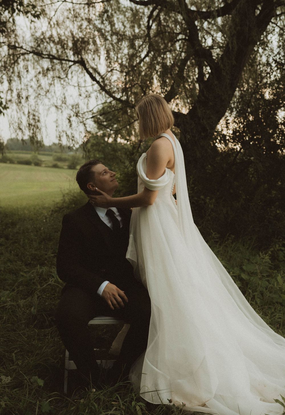 Bride in white gown and groom in black suit share romantic moment under willow tree at golden hour.